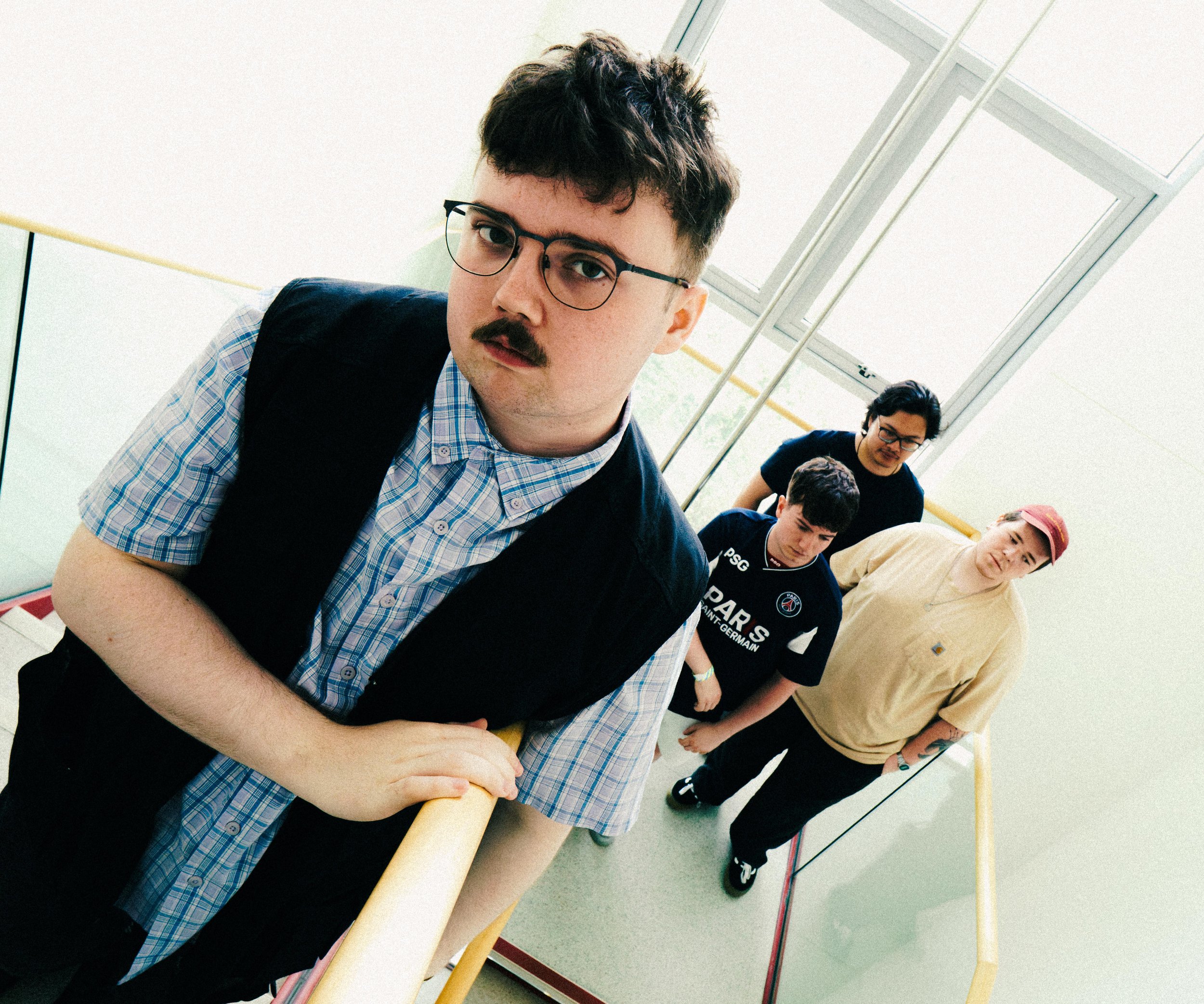 Group of young men standing in a line against a yellow handrail in a bright room with large windows, some with glasses, wearing casual and sporty clothing.