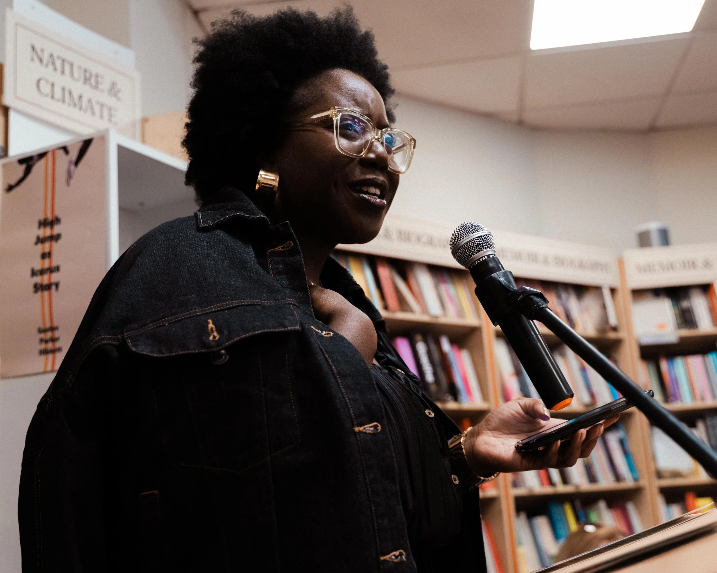 A woman with dark skin, curly hair, wearing glasses, reading or speaking into a microphone in a bookstore or library.