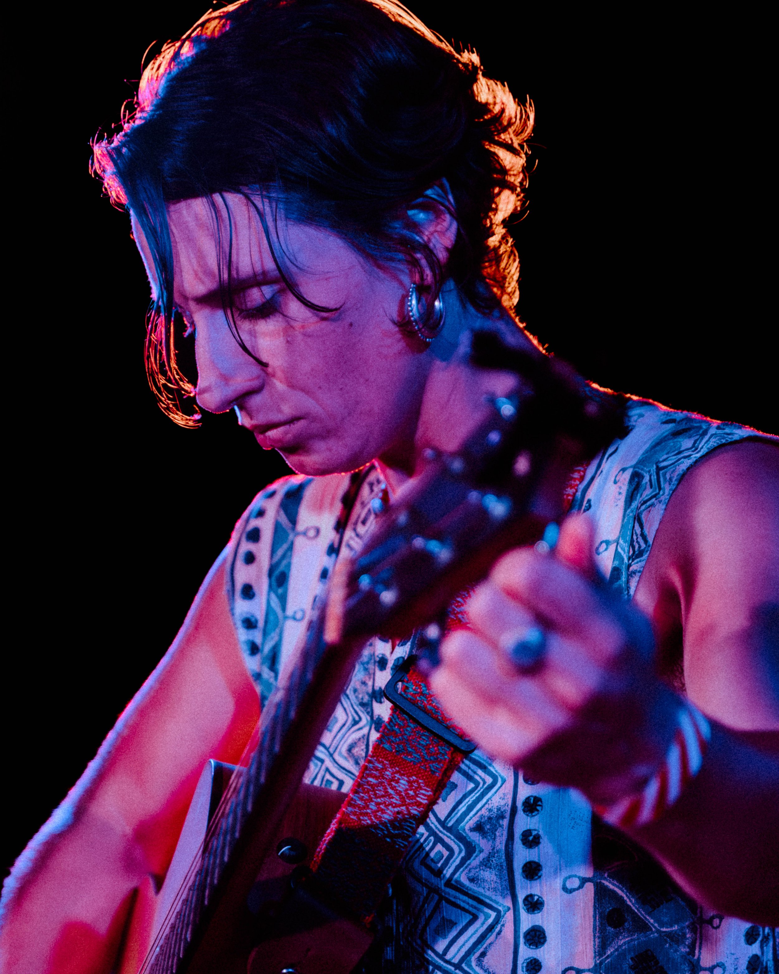 Close-up of a woman with short dark hair and earrings, wearing a patterned shirt, illuminated with colorful lighting against a black background.