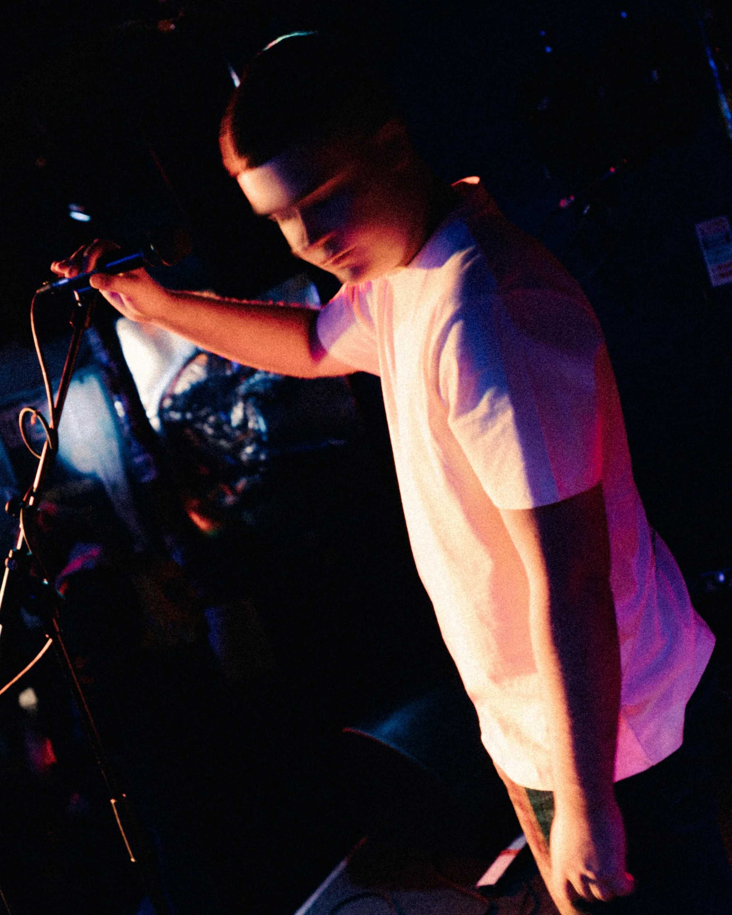 A young person with short black hair and glasses, wearing a white T-shirt, is holding a microphone and appears to be performing or speaking on a dark stage with colorful lighting.