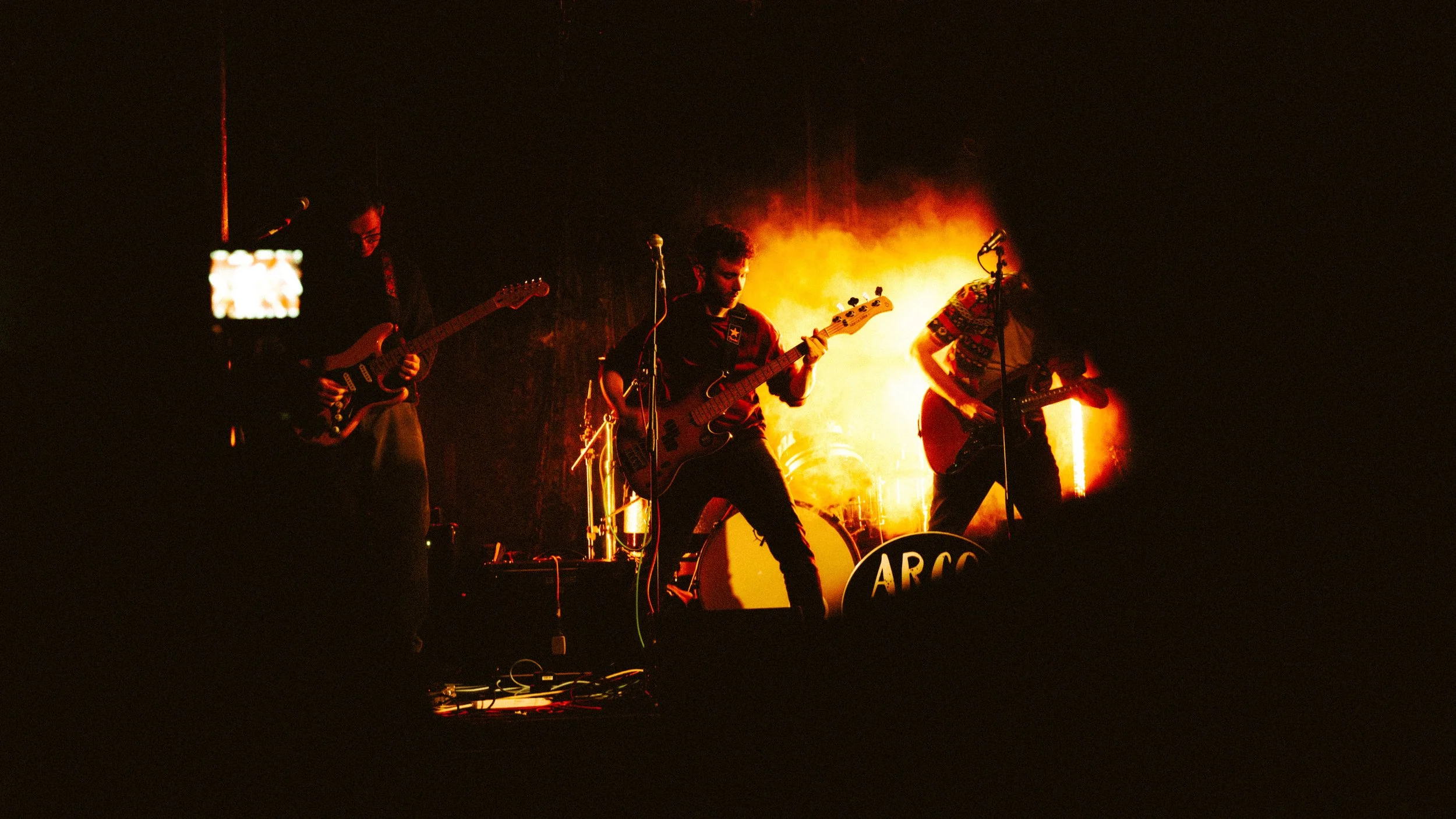 Band performing on stage with orange lighting and smoke effects, featuring three guitar players and a drummer.