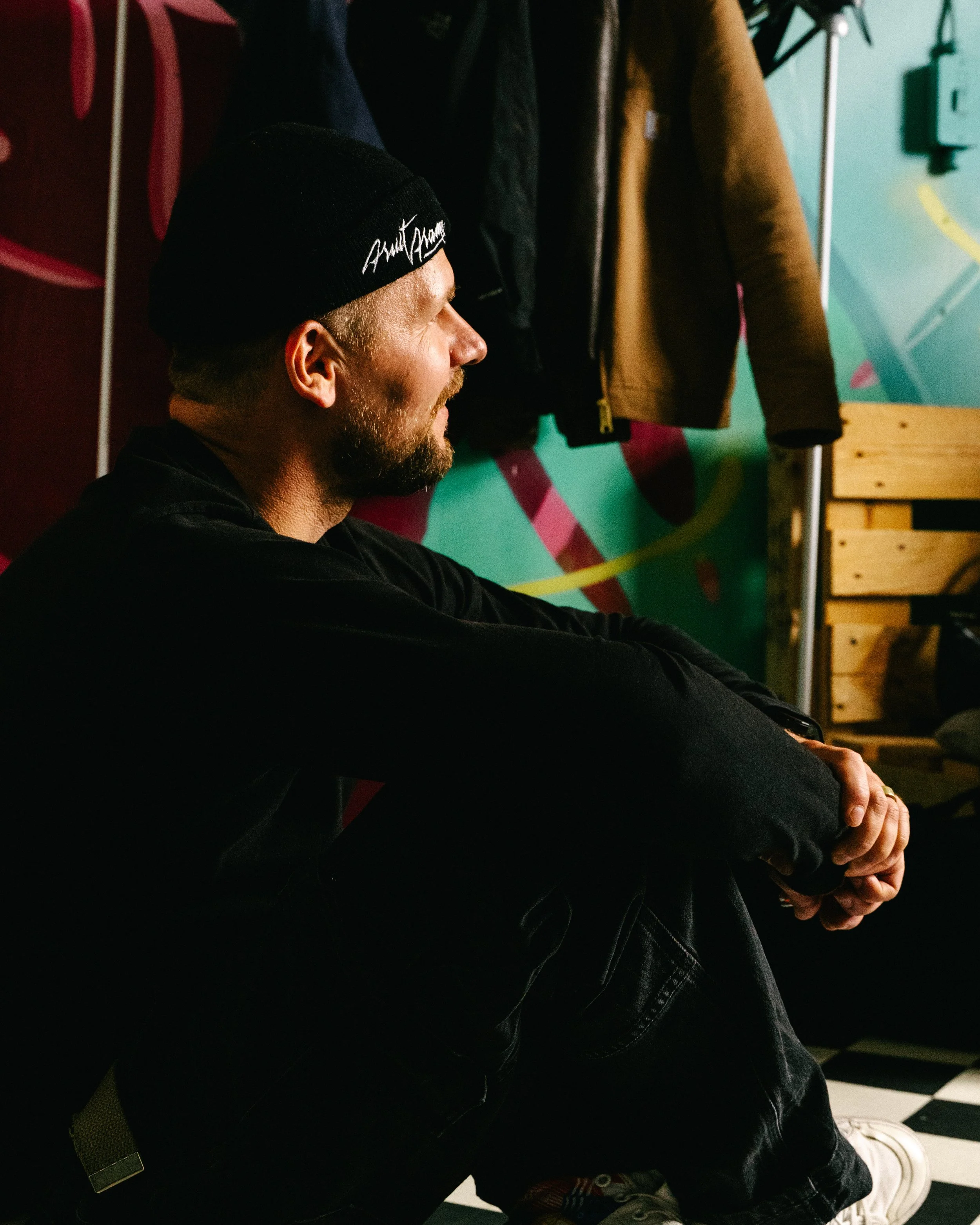 A man sitting with a beanie hat, leaning back with his hands clasped, in a room with colorful wall art and hanging jackets.