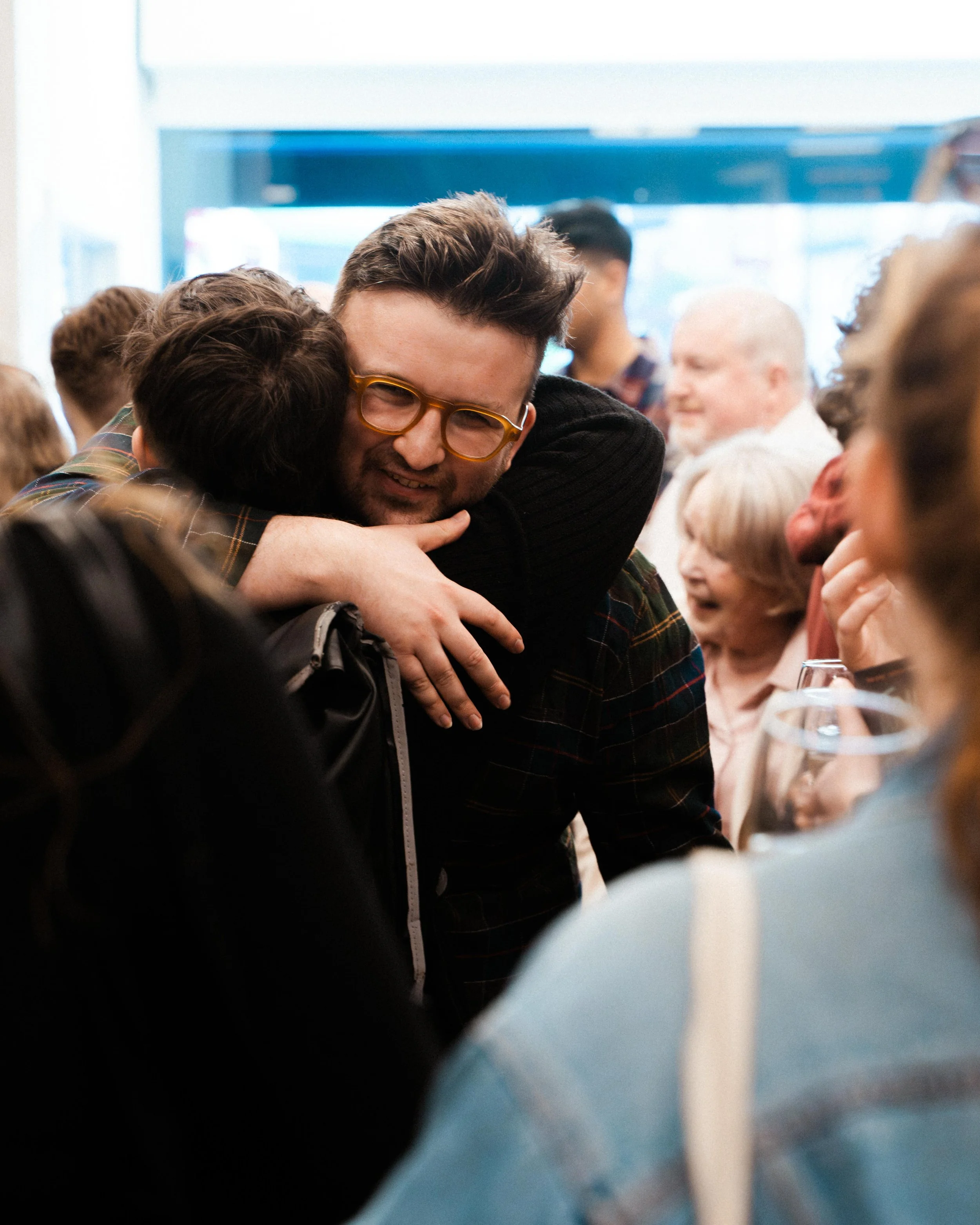 People in a social gathering hugging each other, with several others smiling and engaging in conversation in the background.