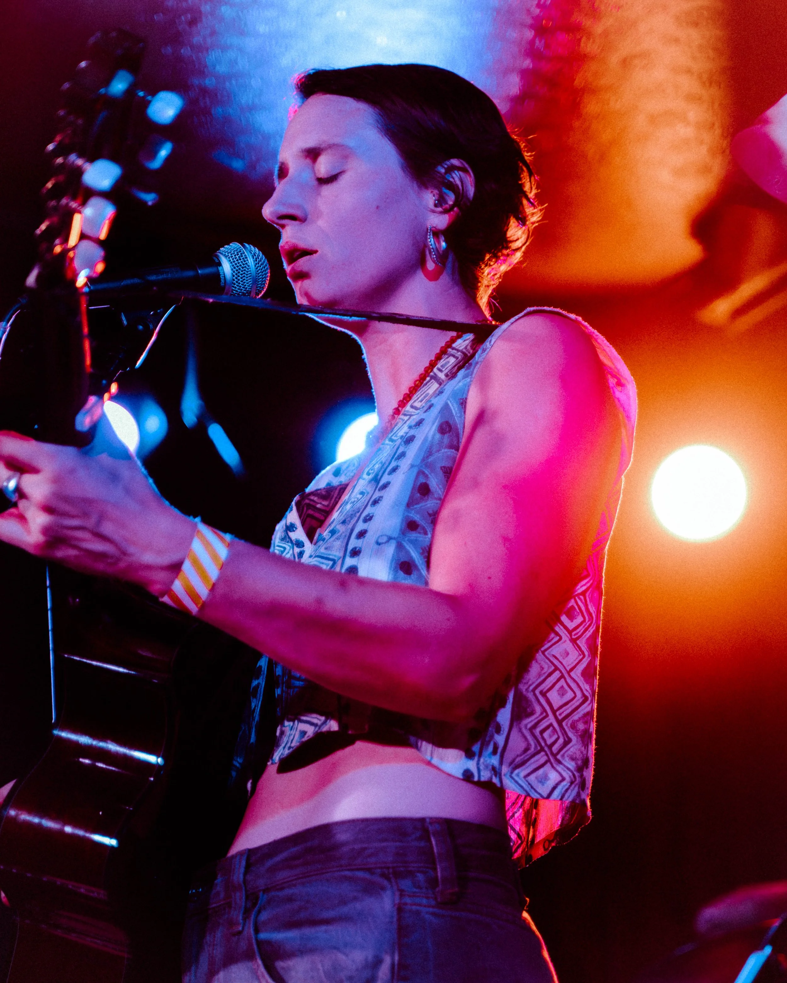 A woman with short dark hair is singing into a microphone while playing an acoustic guitar. She is wearing earrings, a sleeveless top, and is illuminated by colorful stage lighting, with a blurred background.