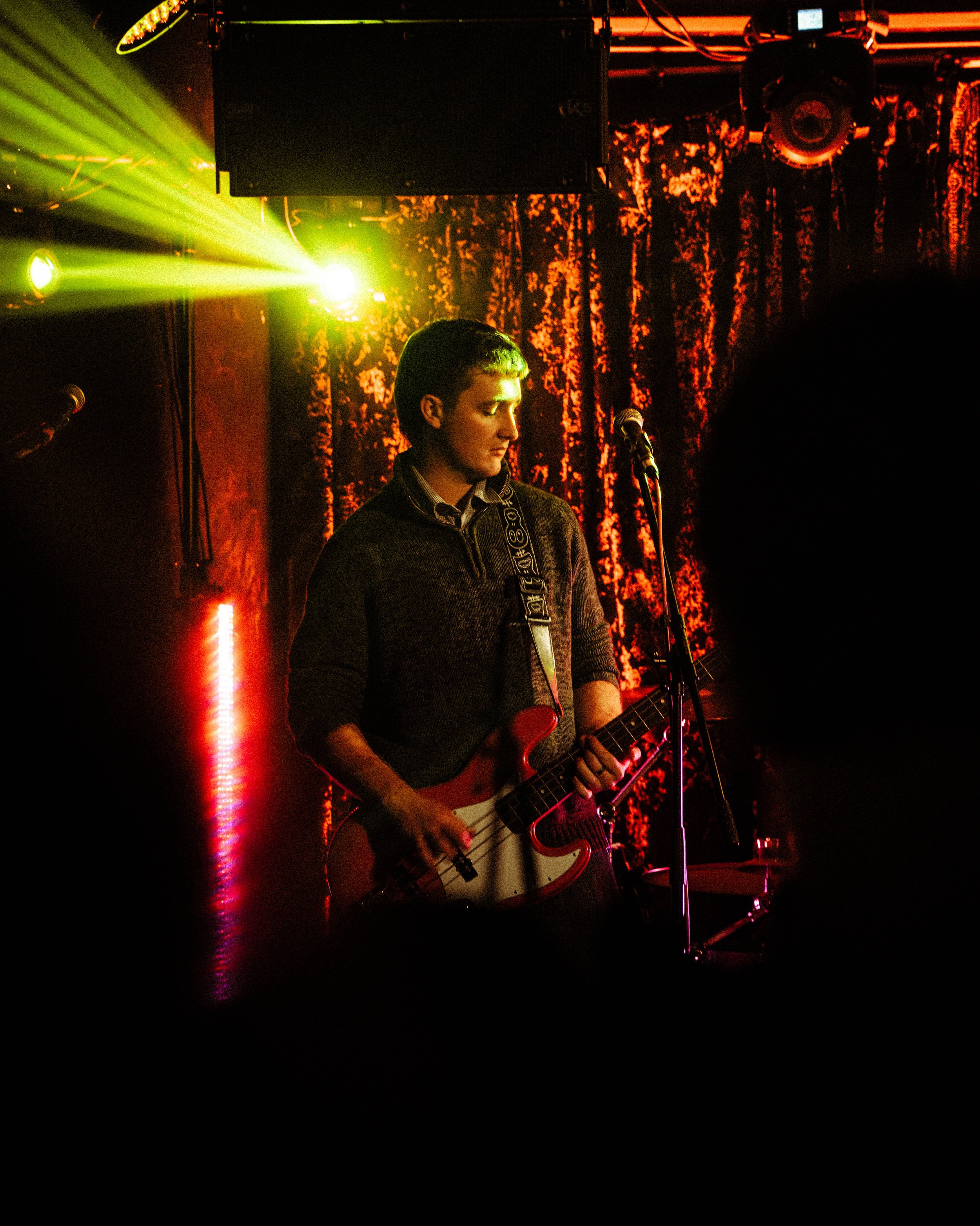 A young man playing an electric guitar on stage during a live music performance, illuminated by colorful stage lighting with a backdrop of textured curtains.