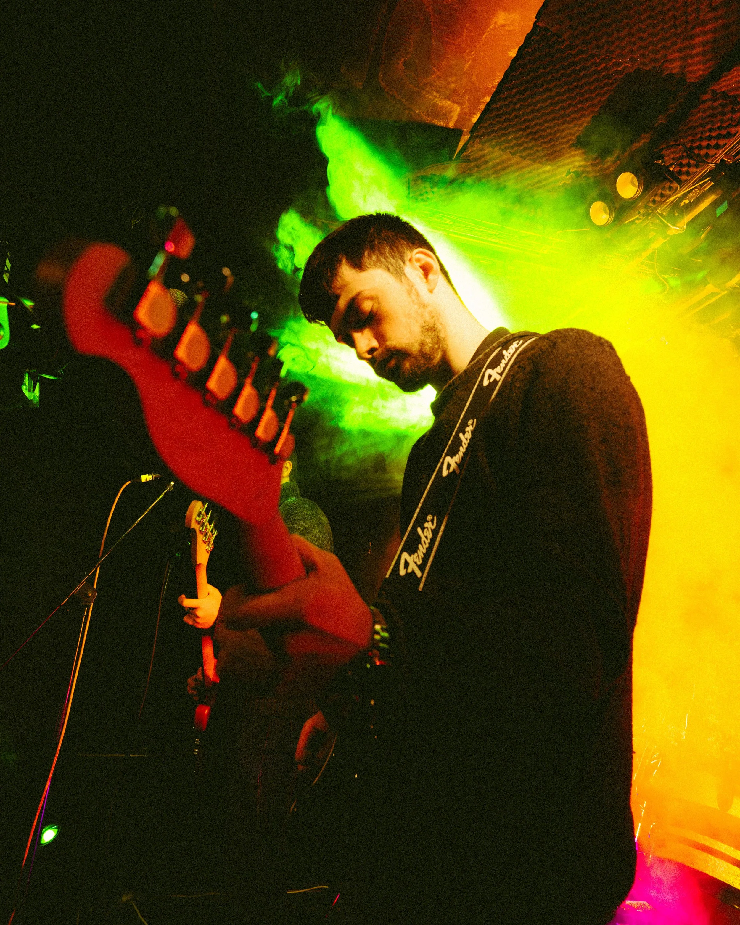 A male musician playing an electric guitar on stage with colorful stage lighting, including green and yellow lights, in a dark venue.