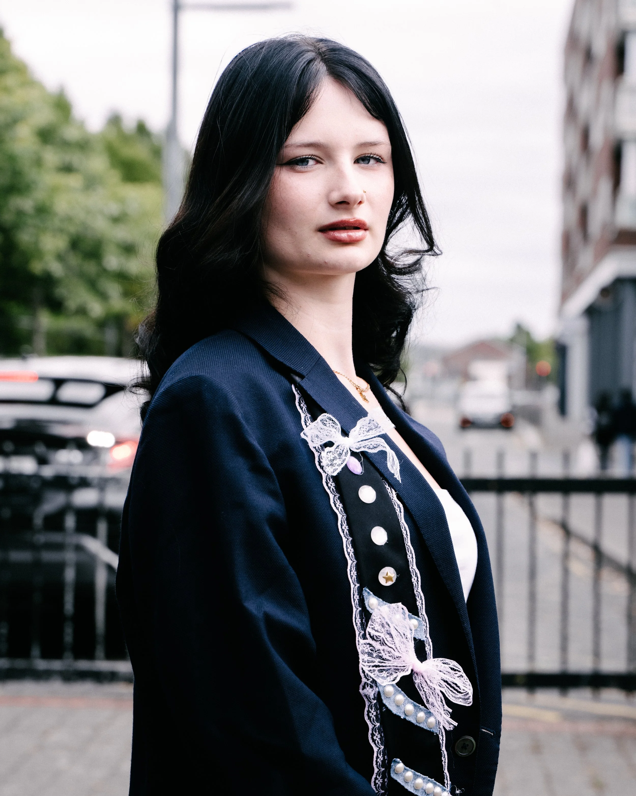 A woman with black hair and blue eyes wearing a black and white outfit with lace and pearl details, standing outdoors with blurred cars and trees in the background.