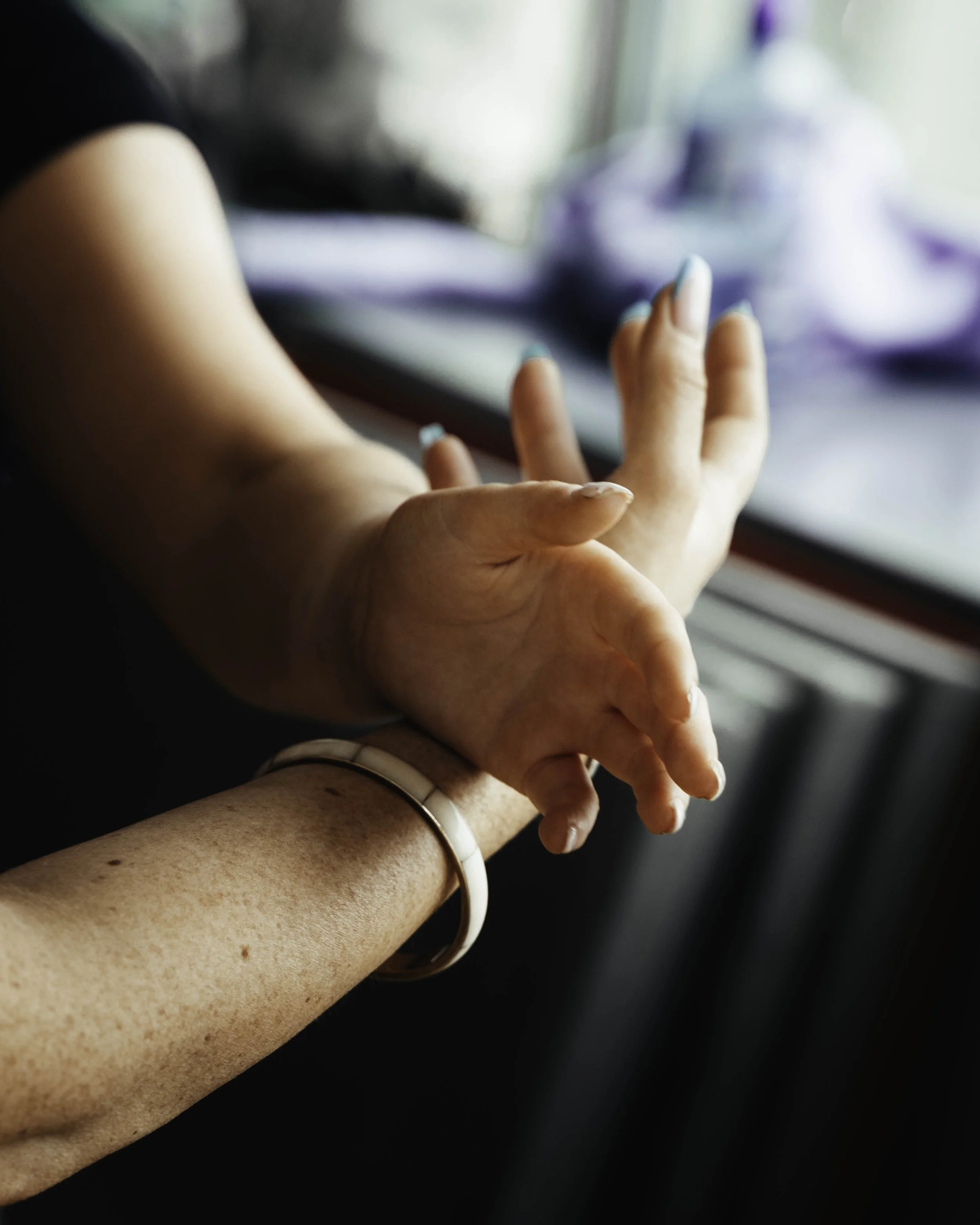 Close-up of two hands gently clasped, with a blurred background. One hand has painted nails and a bracelet.