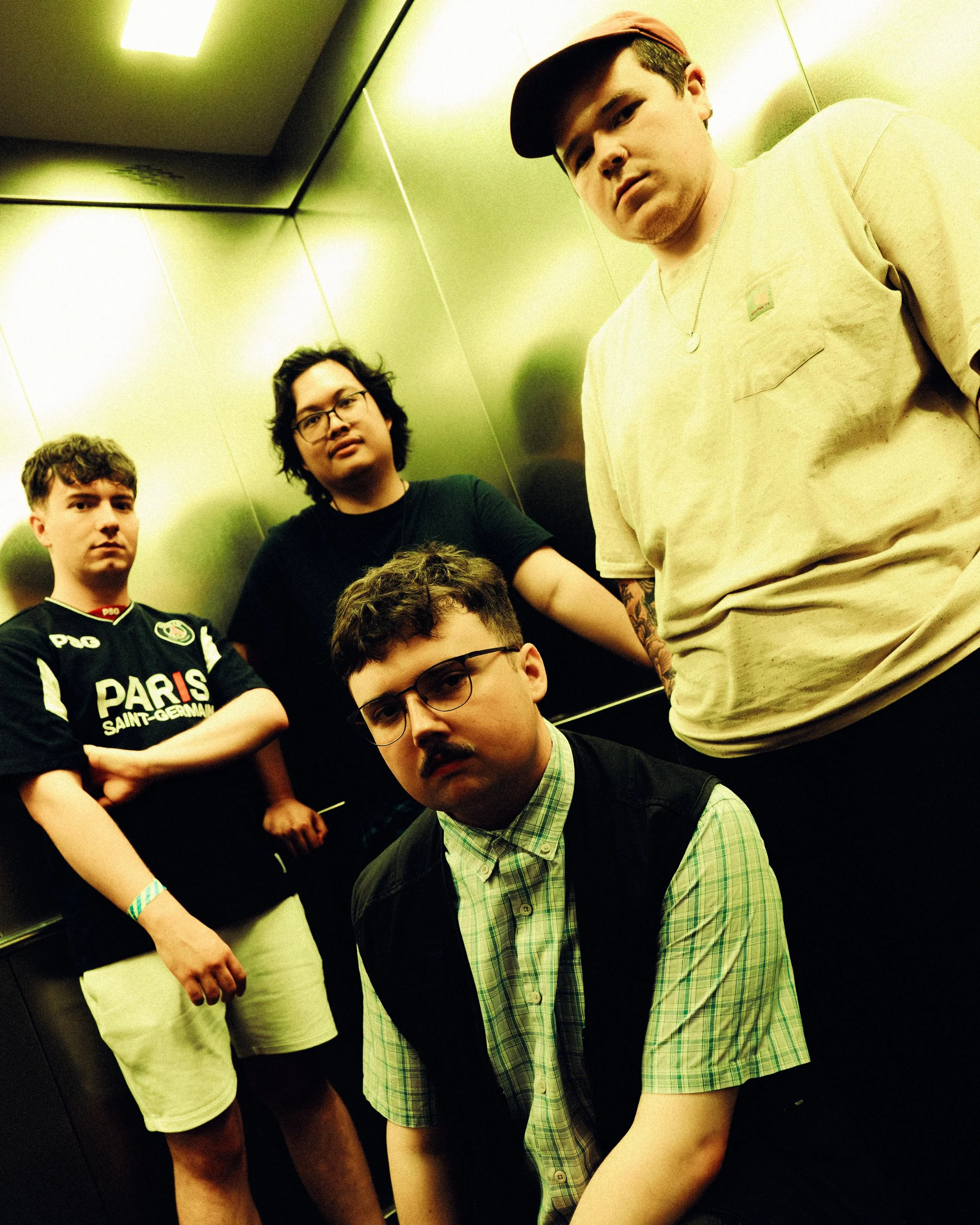 Group of young men in an elevator posing for a photo. One person in the foreground with glasses and a mustache, wearing a green plaid shirt and a black vest. Others in the background wearing casual clothes, including one with a black sports jersey an