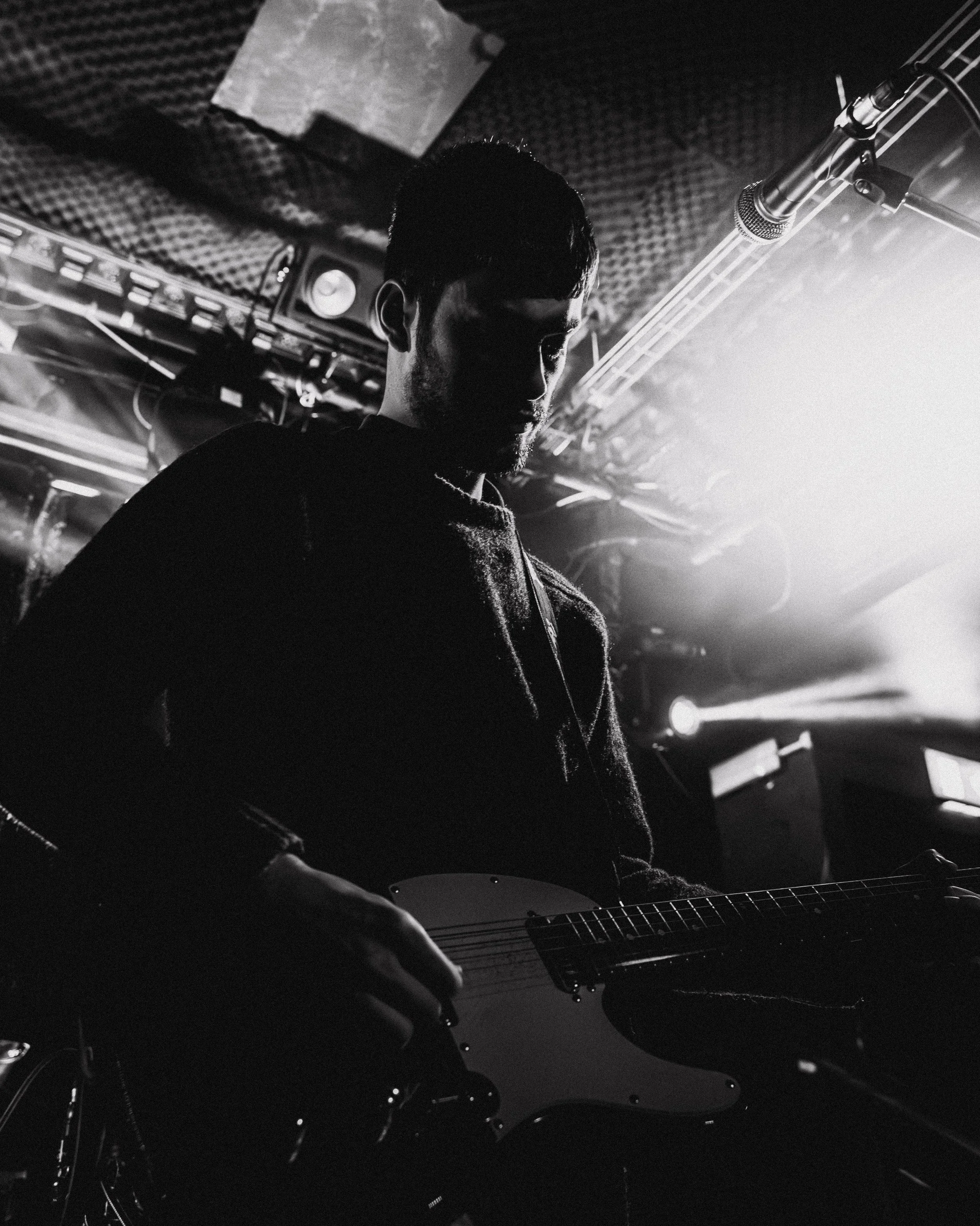 A silhouette of a man playing an electric guitar in a dimly lit studio or stage with bright backlighting.