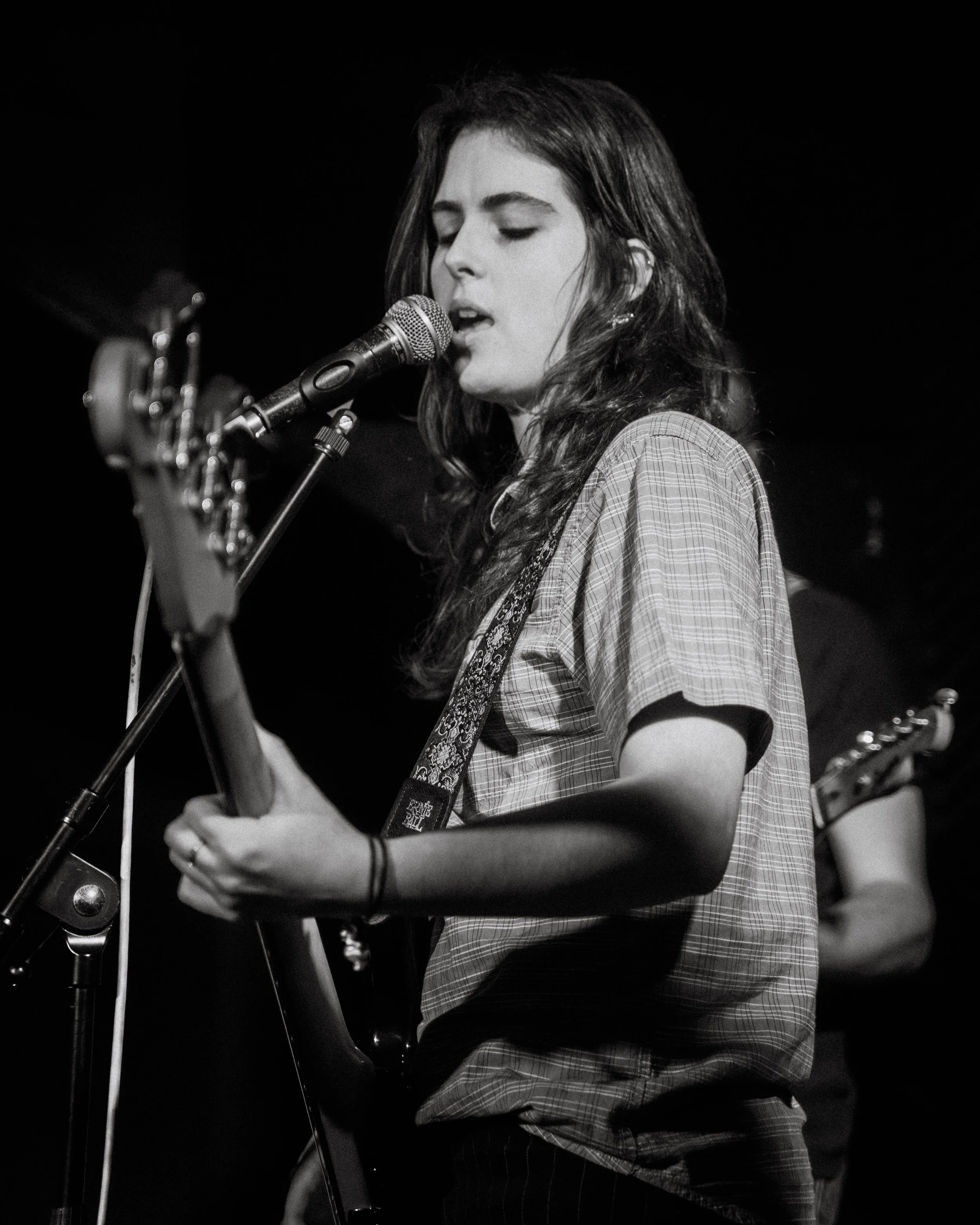 A young woman with long hair sings into a microphone while playing an electric guitar during a live music performance.