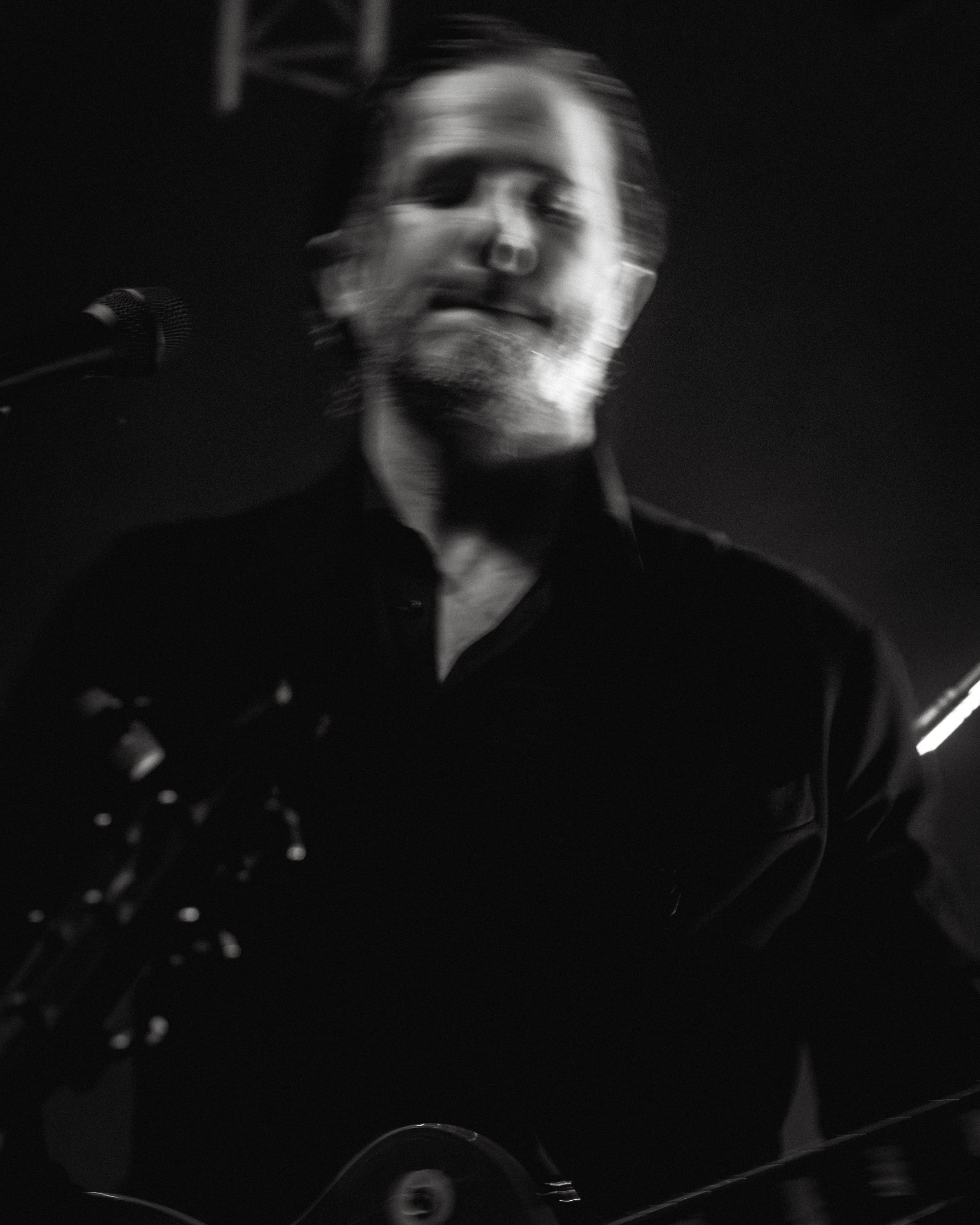 A black and white photo of a man playing guitar and singing into a microphone on stage, with a blurred, motion effect.
