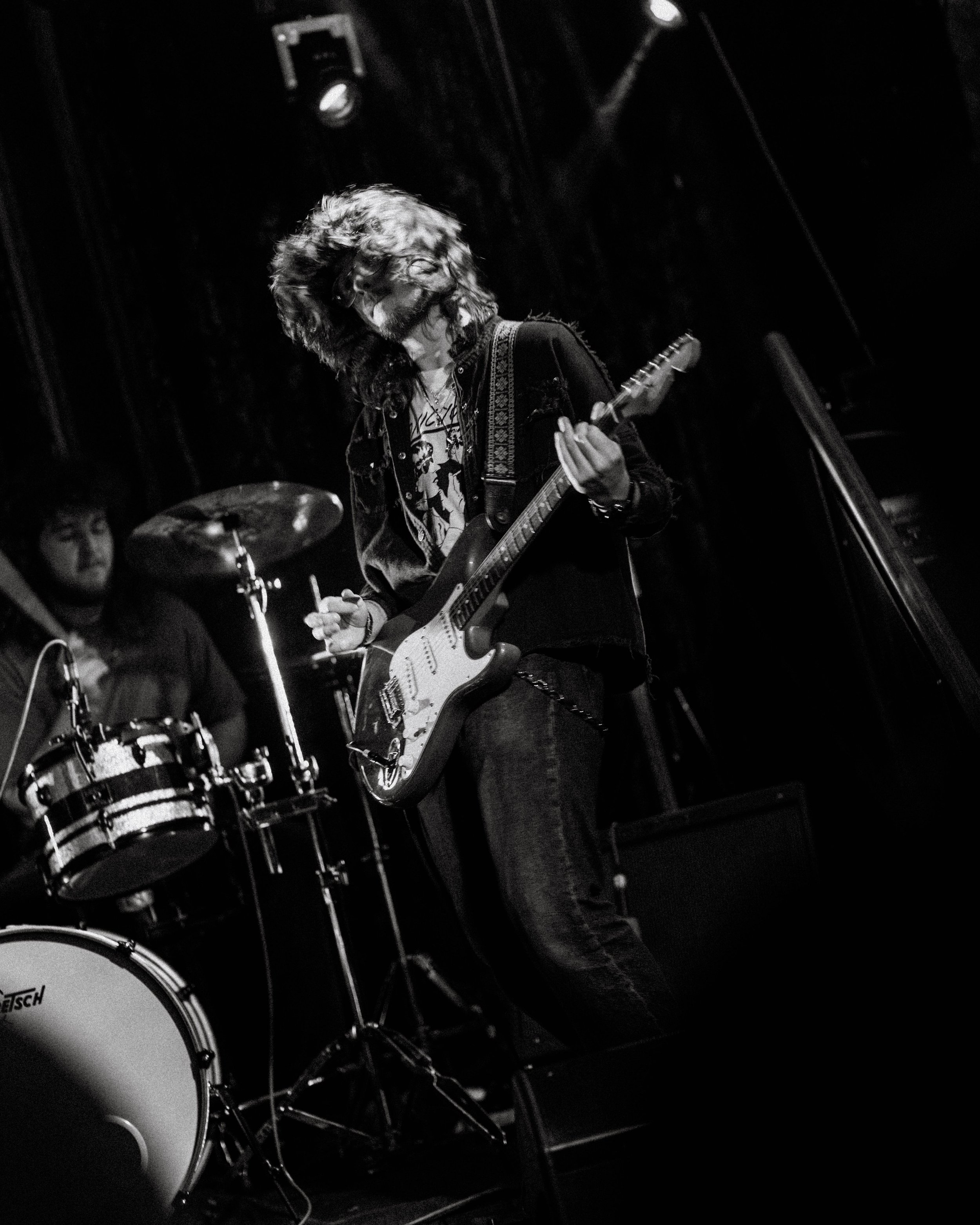 A black and white photo of a musician playing an electric guitar with long, curly hair, with a drummer in the background, on stage.