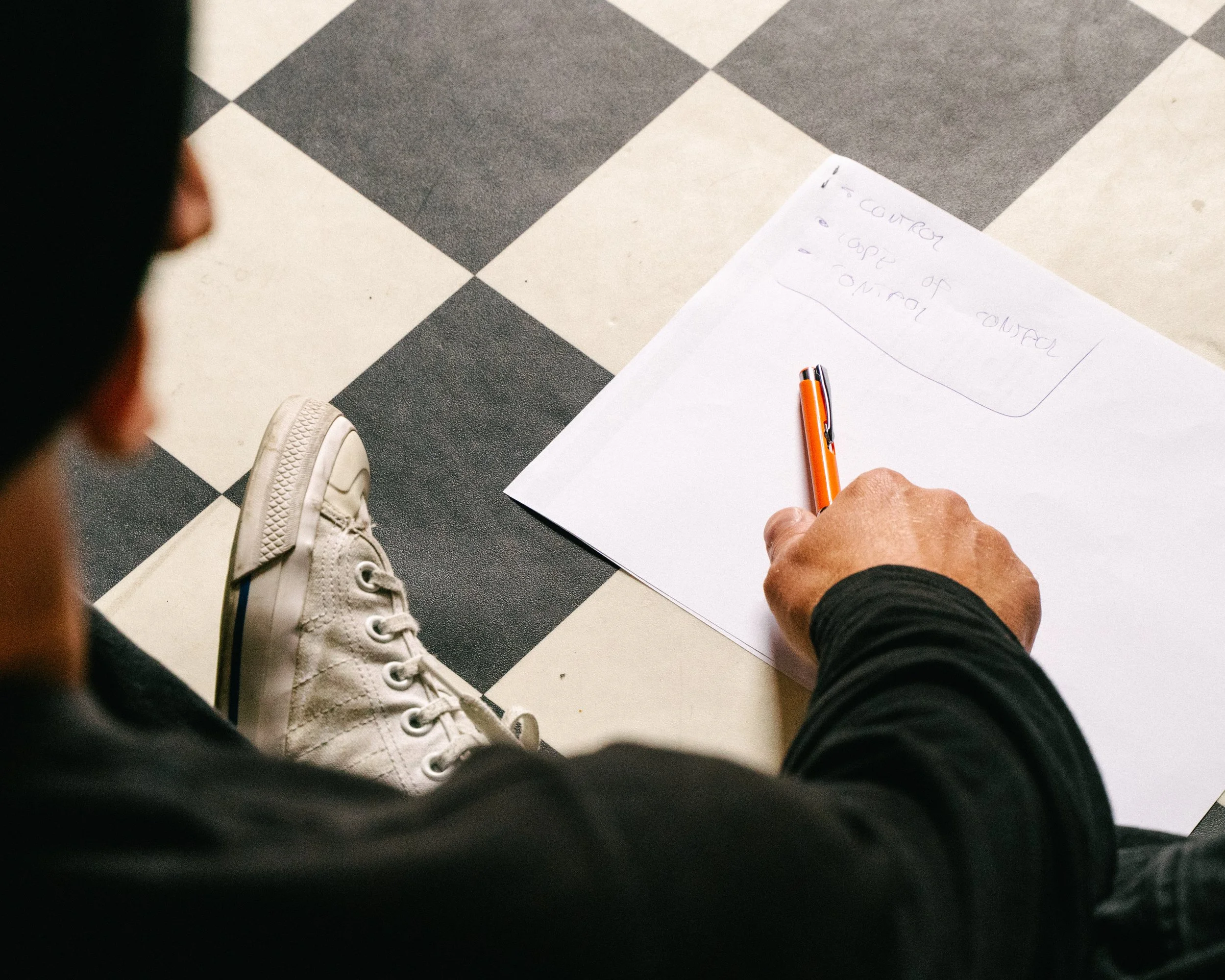 A person sitting on the floor, wearing white sneakers and black pants, writing on a white piece of paper with an orange pen. The paper has some handwritten notes. The area has black and white checkered flooring.