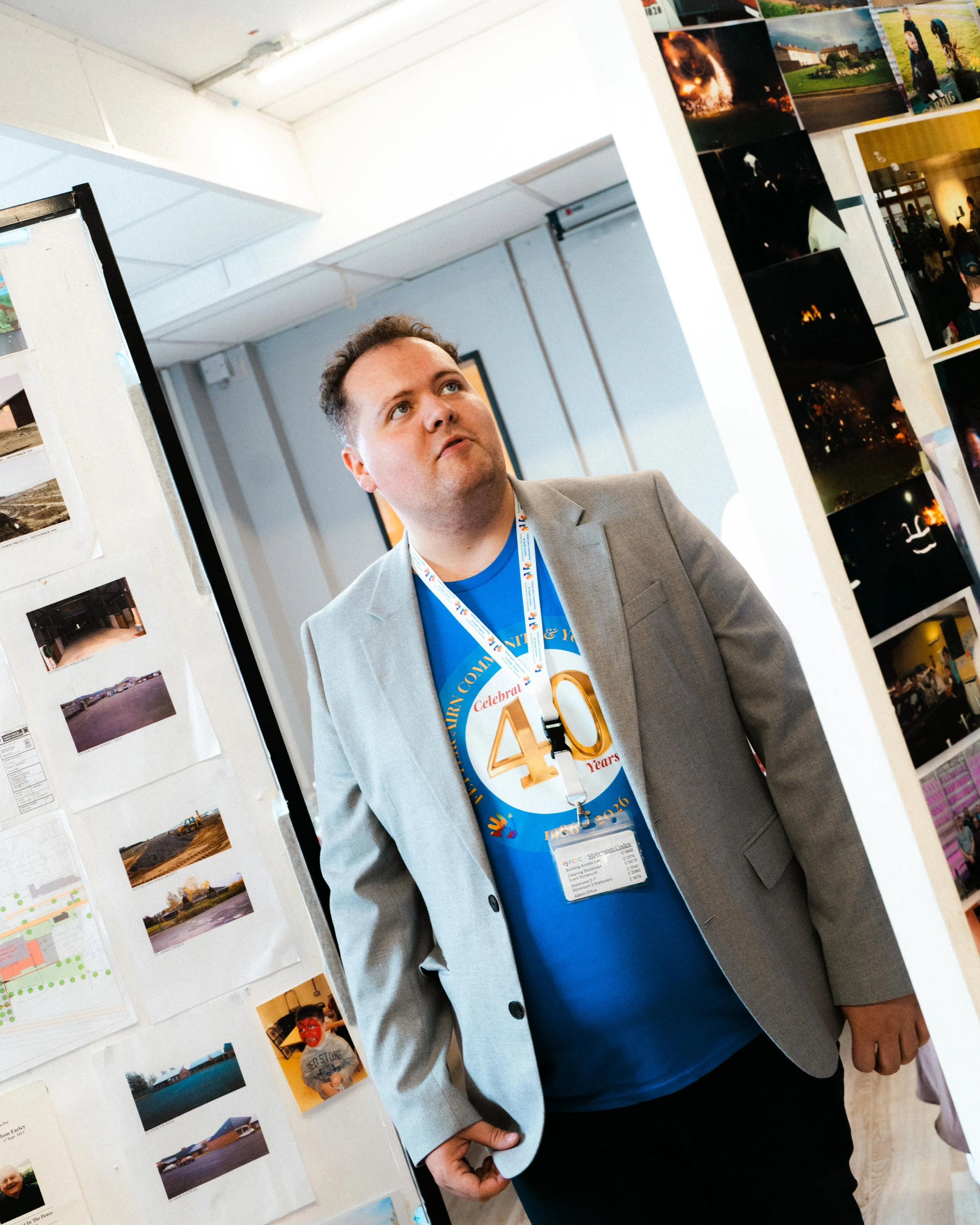 A man in a gray blazer and blue shirt standing in front of a photo display at an event. He is wearing a commemorative medal on a lanyard around his neck, which reads 'Celebrating 40 Years'. The display features photographs and images attached to whit