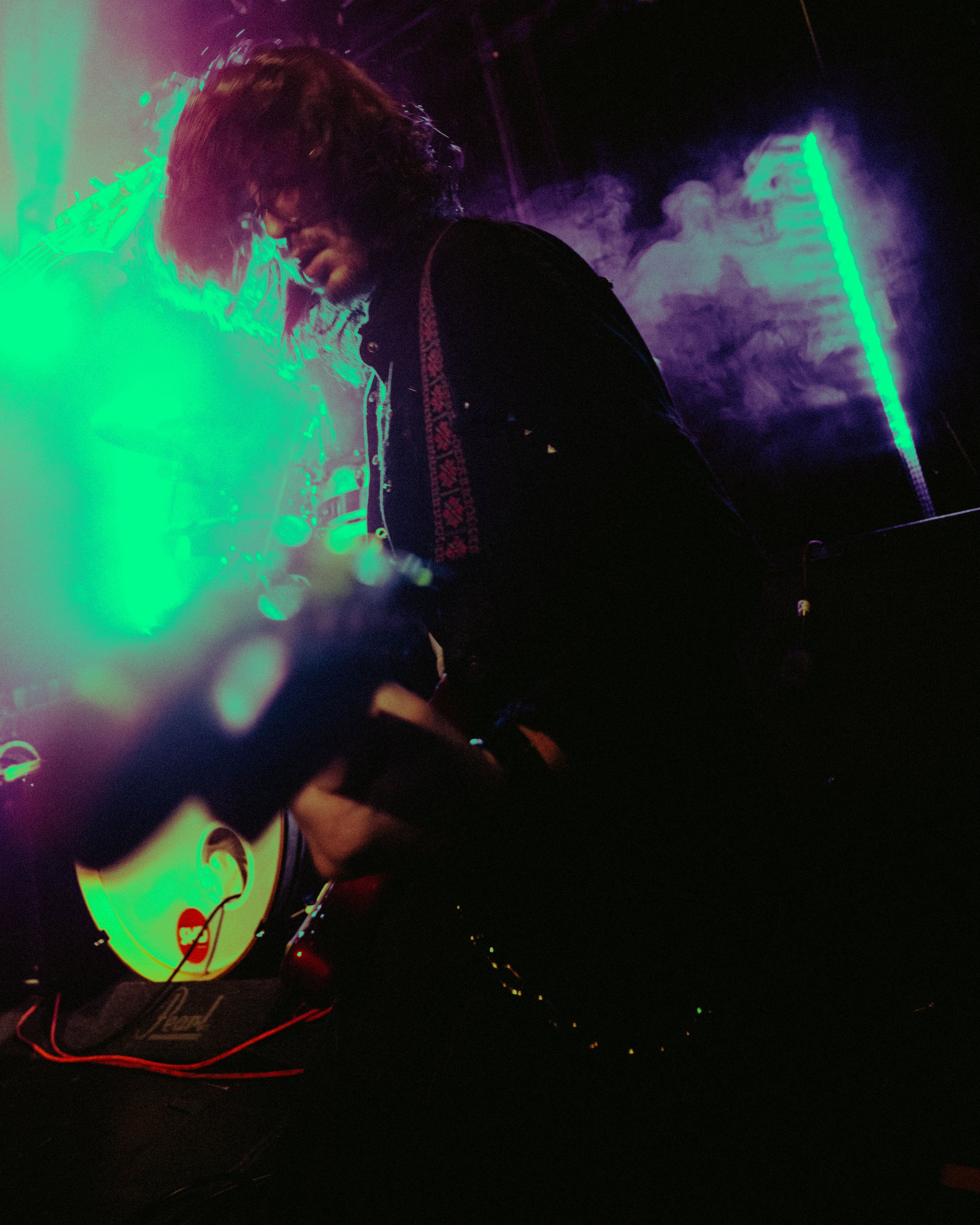 Person with long hair playing guitar on stage with colorful neon lights and smoke effects.