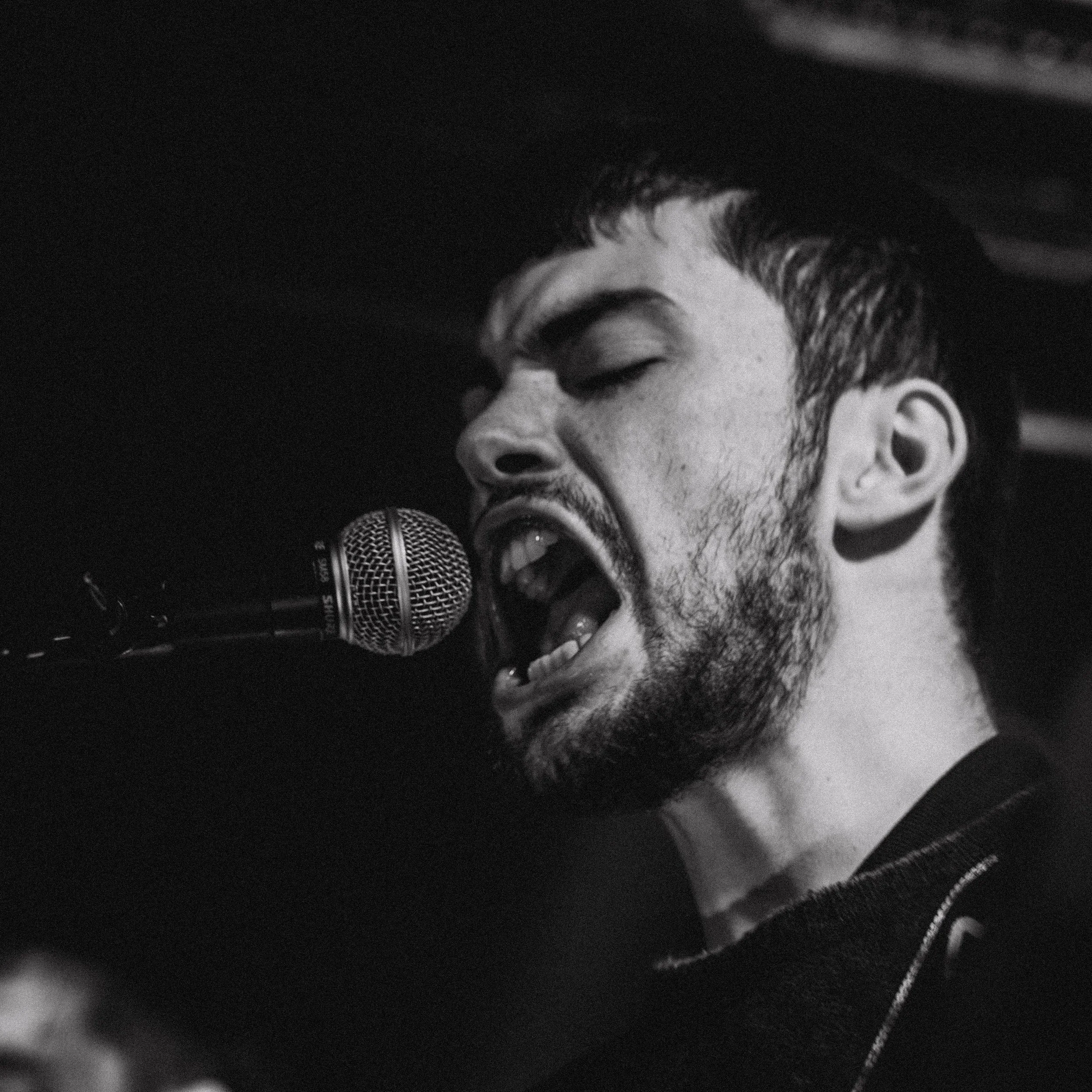 A black and white photo of a man singing passionately into a microphone, with eyes closed and mouth wide open.