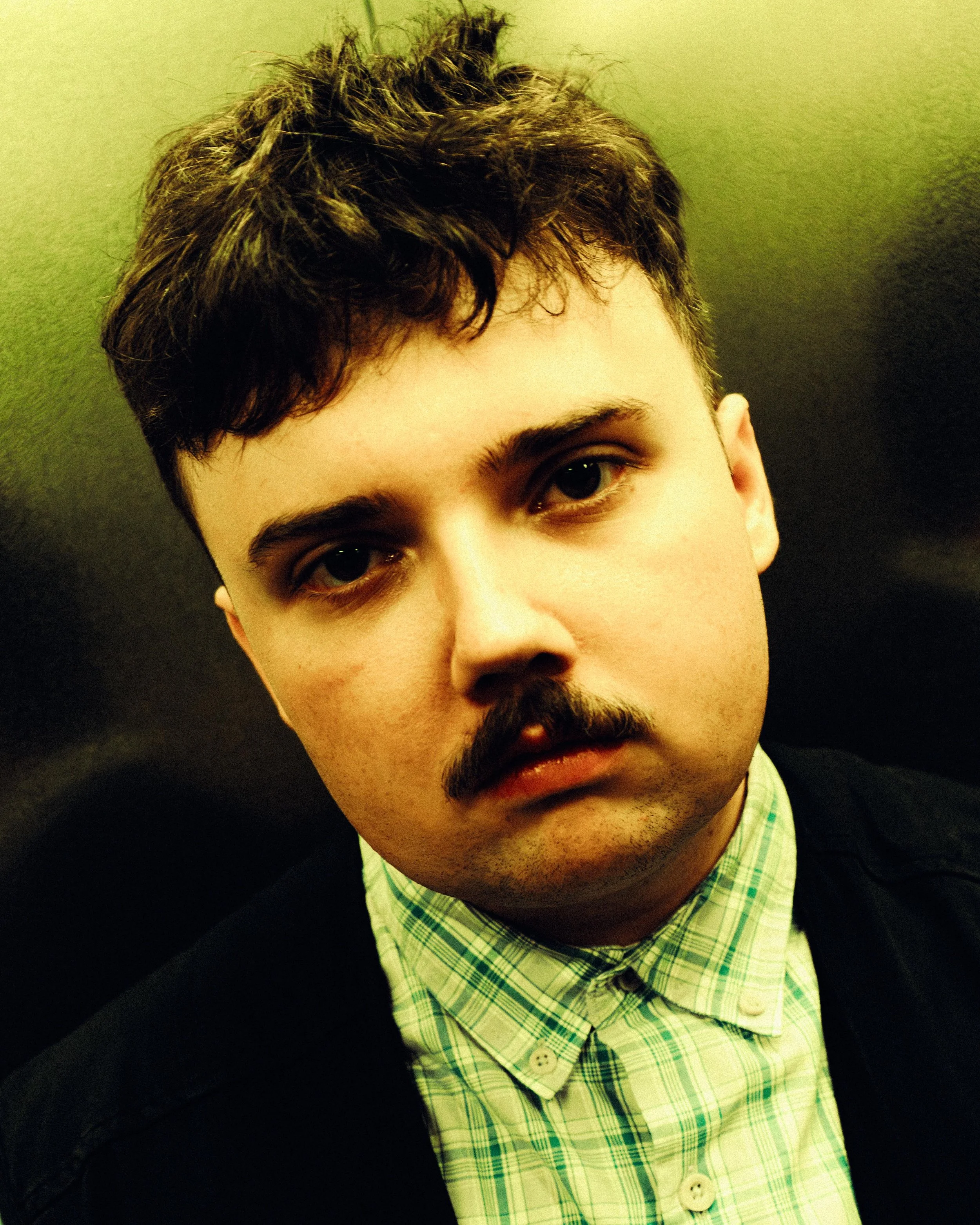 Close-up of a person's face with curly hair, brown eyes, and a mustache, wearing a green plaid shirt and a black jacket, against a black background.