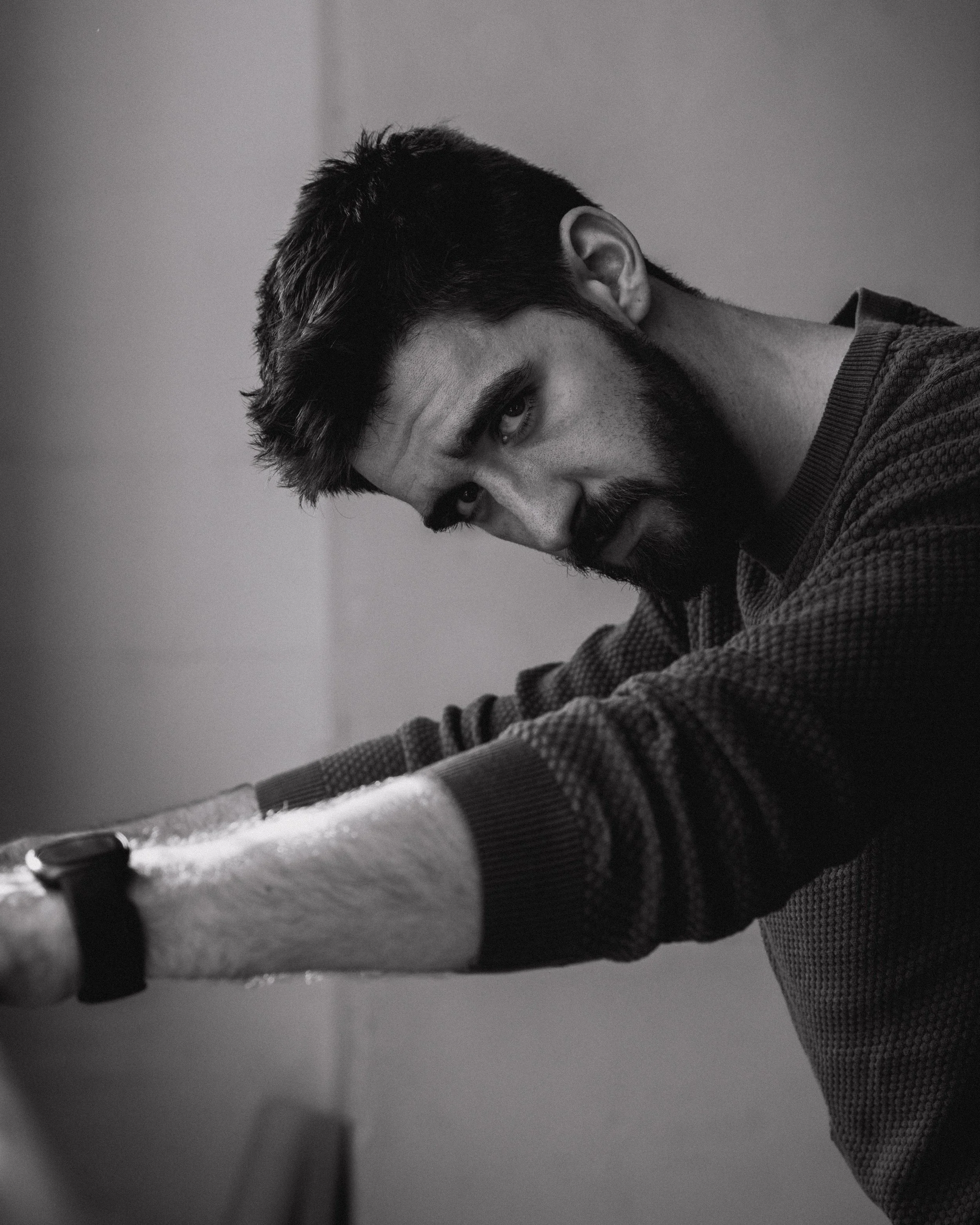 Black and white close-up photo of a young man with dark hair and beard, resting his head on his arm, looking into the camera with a serious expression.