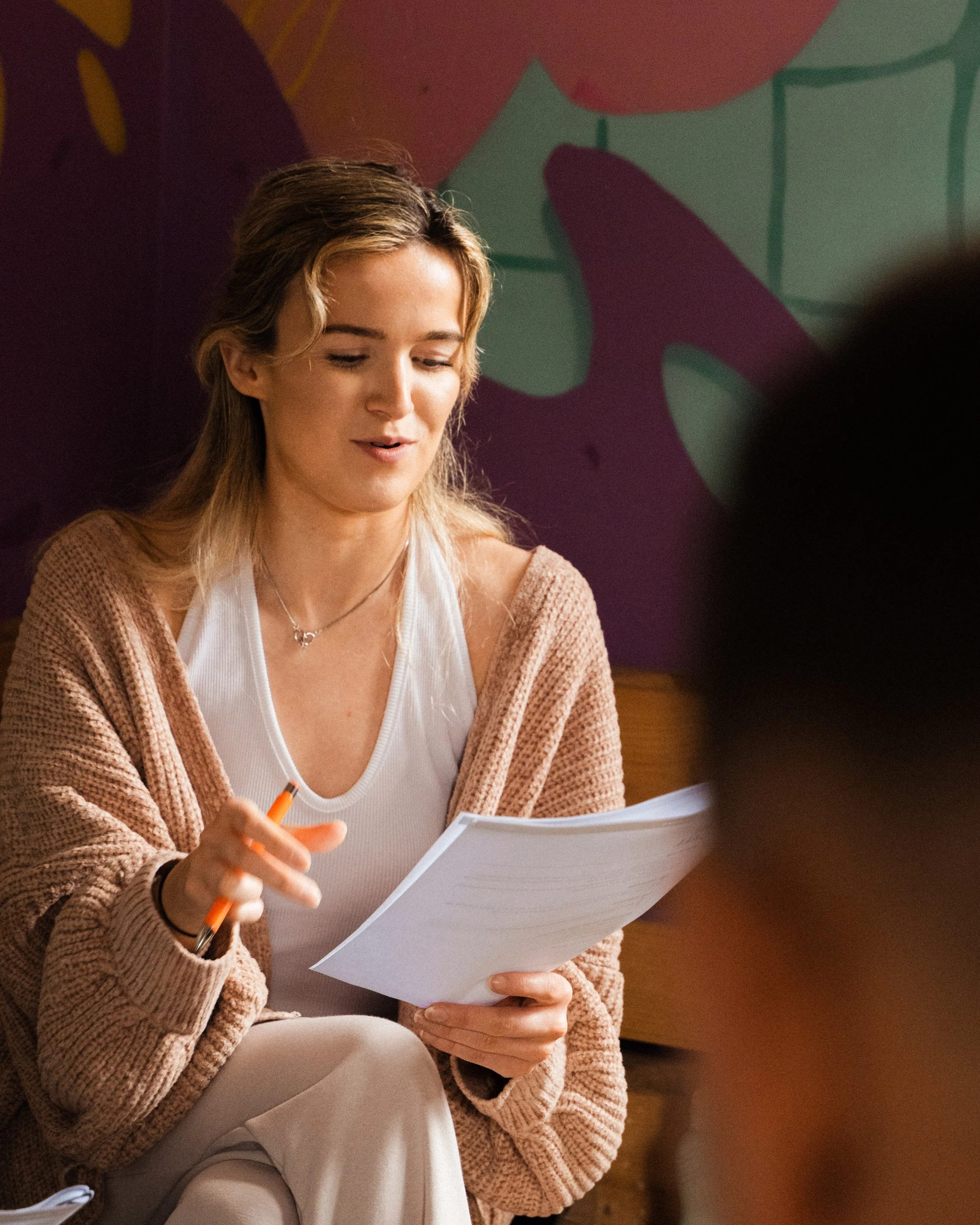 A woman with wavy hair is sitting and holding a piece of paper and an orange pen, wearing a white top and a beige cardigan, with a colorful abstract wall in the background.