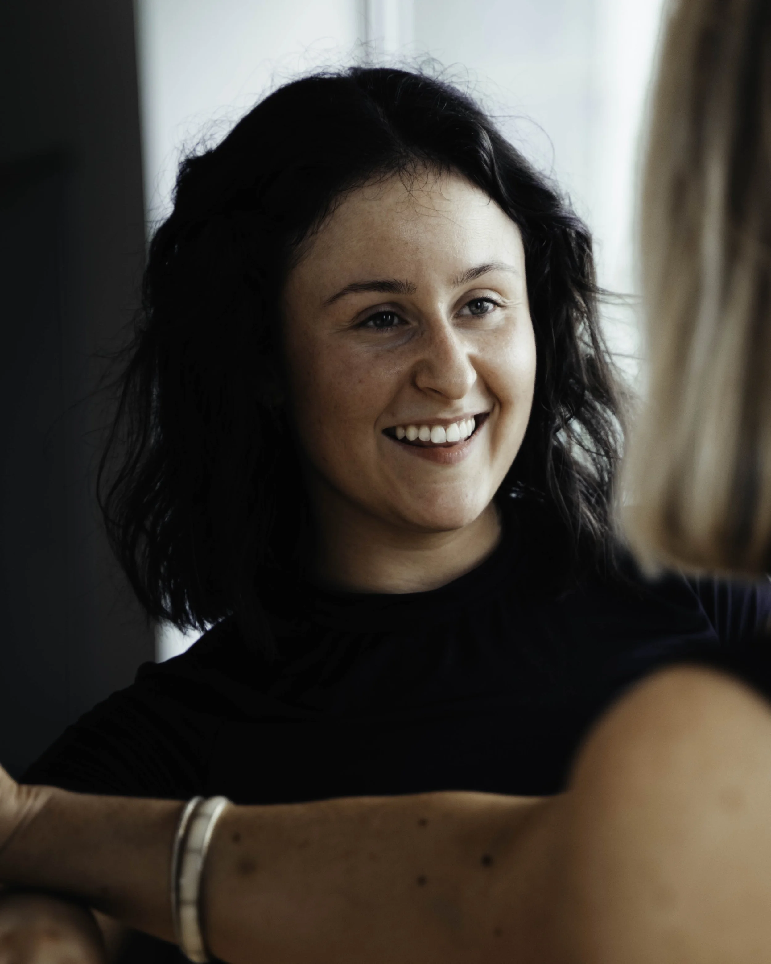 A woman with black, curly hair smiling, wearing a black top and a white bracelet, near a window with curtains.