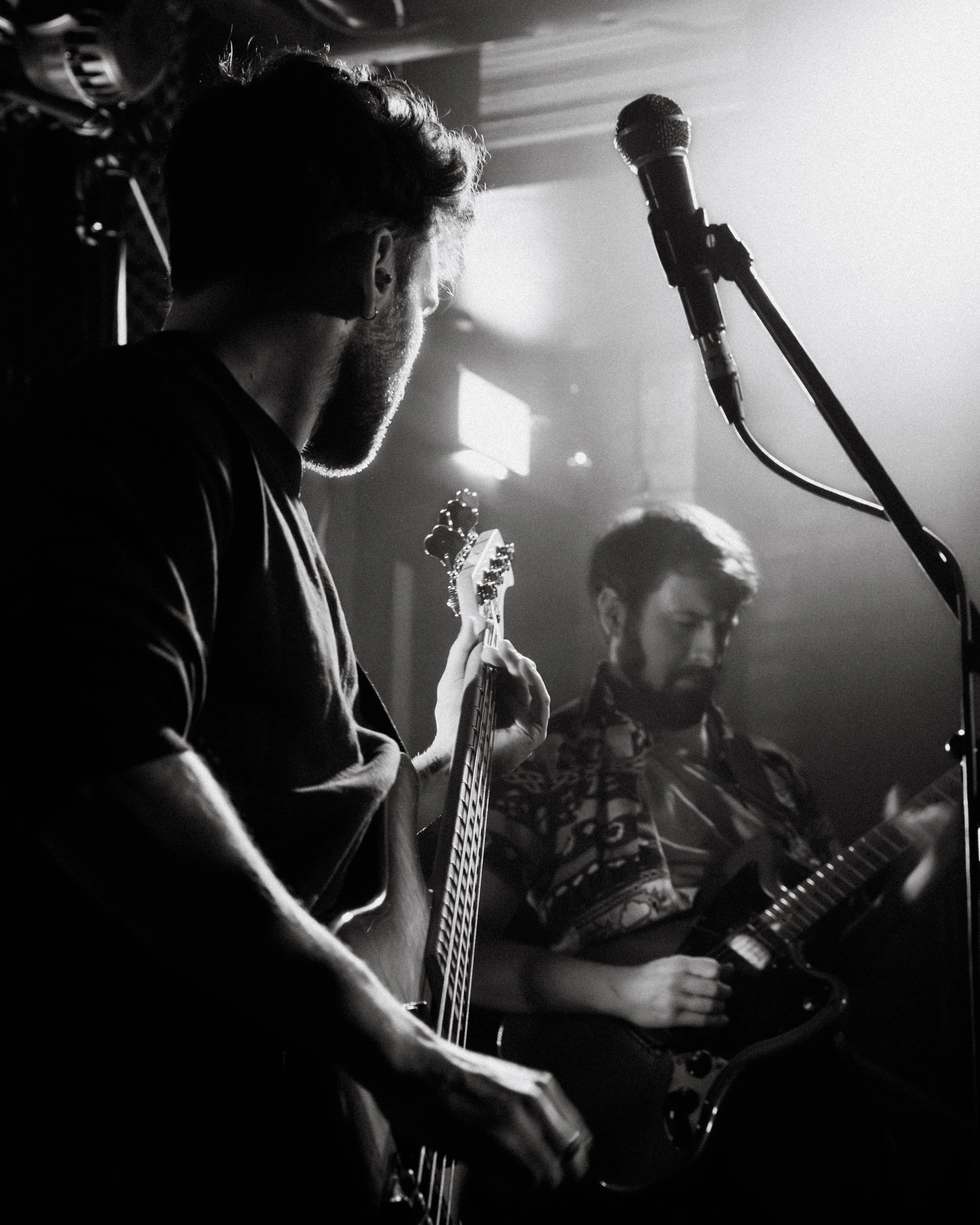 Two musicians playing guitars on stage, with a microphone in the foreground and stage lighting creating a dramatic scene.