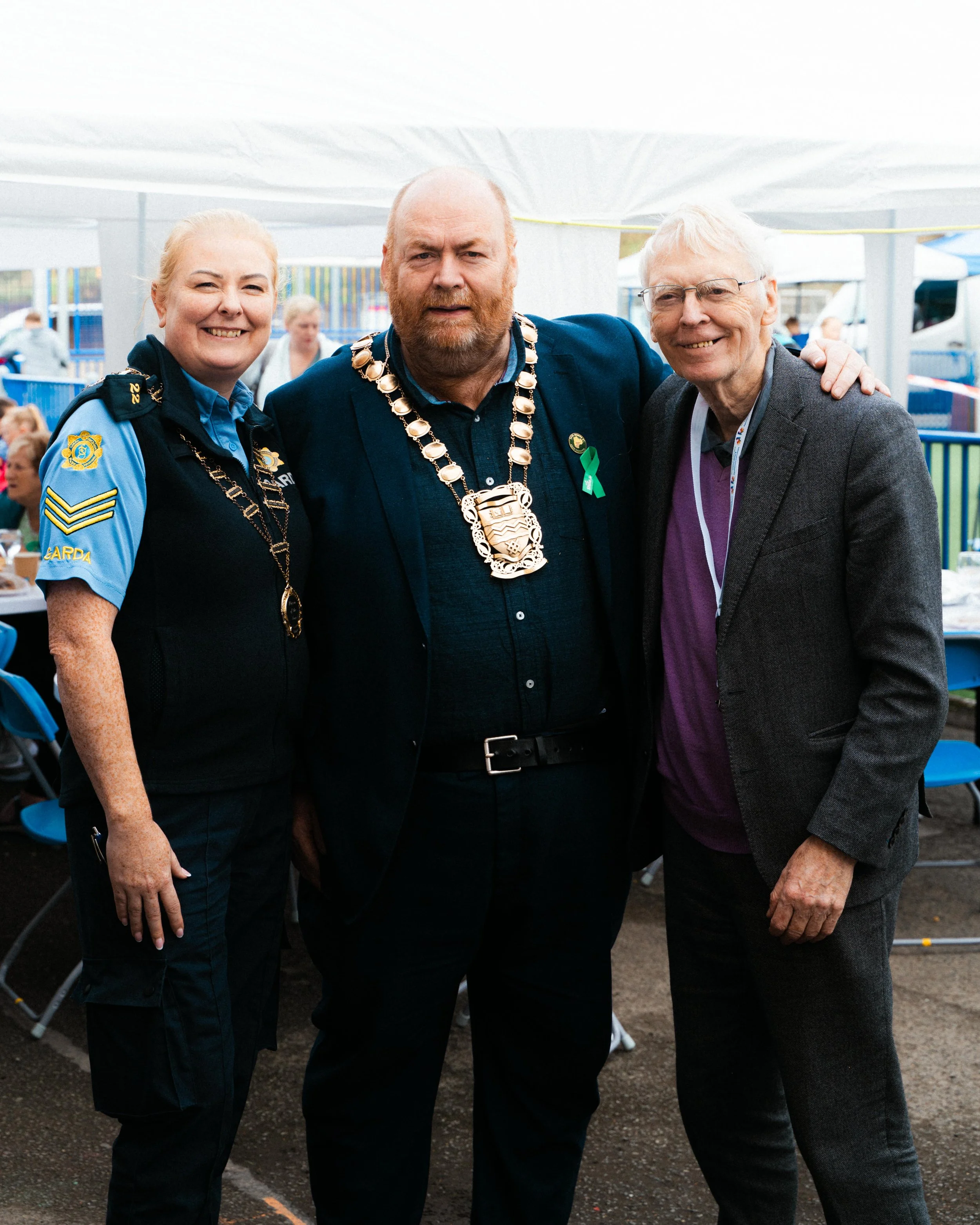 Three people standing together at an outdoor event, with the person in the middle wearing a chain of office, suggesting a ceremonial or formal occasion.