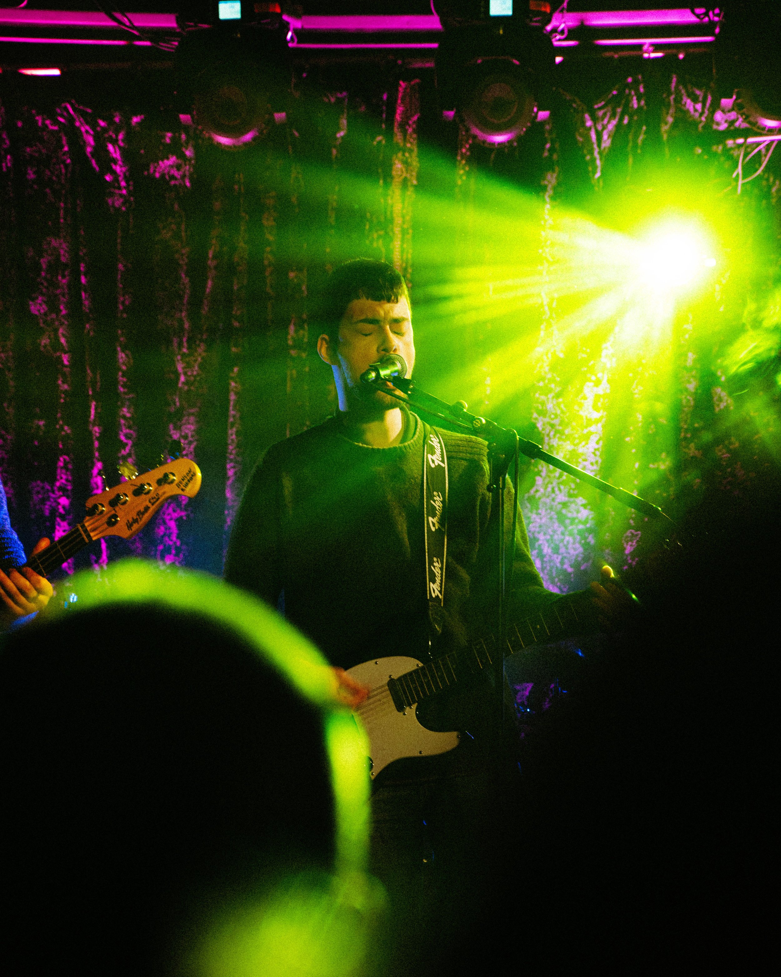 Young male musician singing into a microphone and playing an electric guitar on stage with colorful stage lighting and curtains in the background.