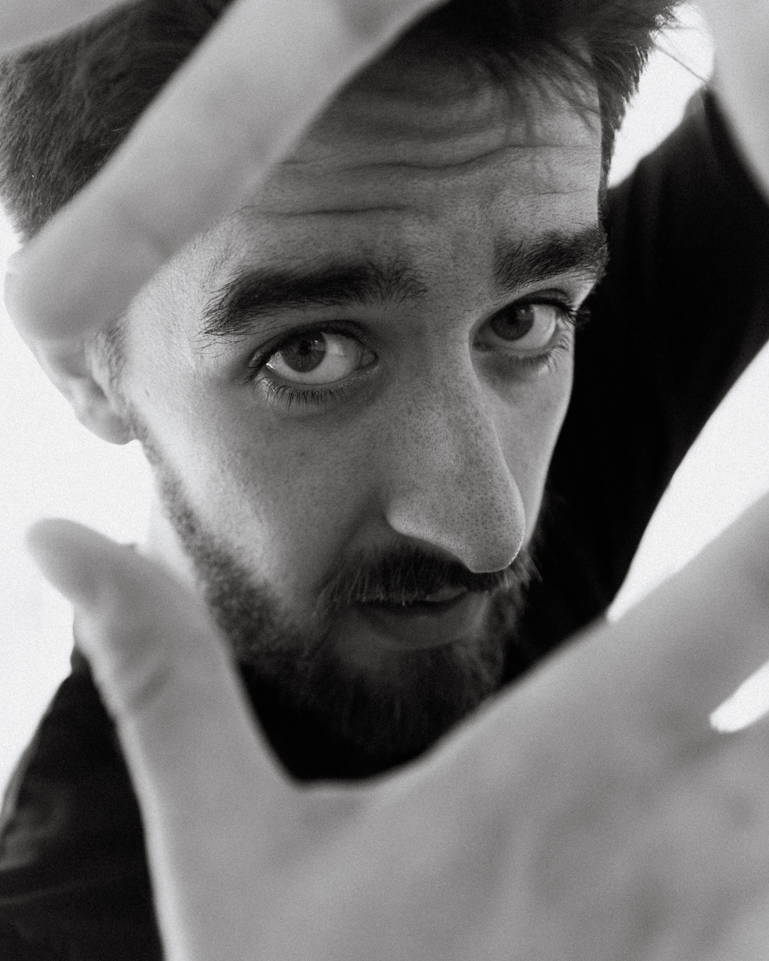 Close-up black and white portrait of a young man with a beard, looking up at the camera through a slightly tilted and circular mirror or lens.