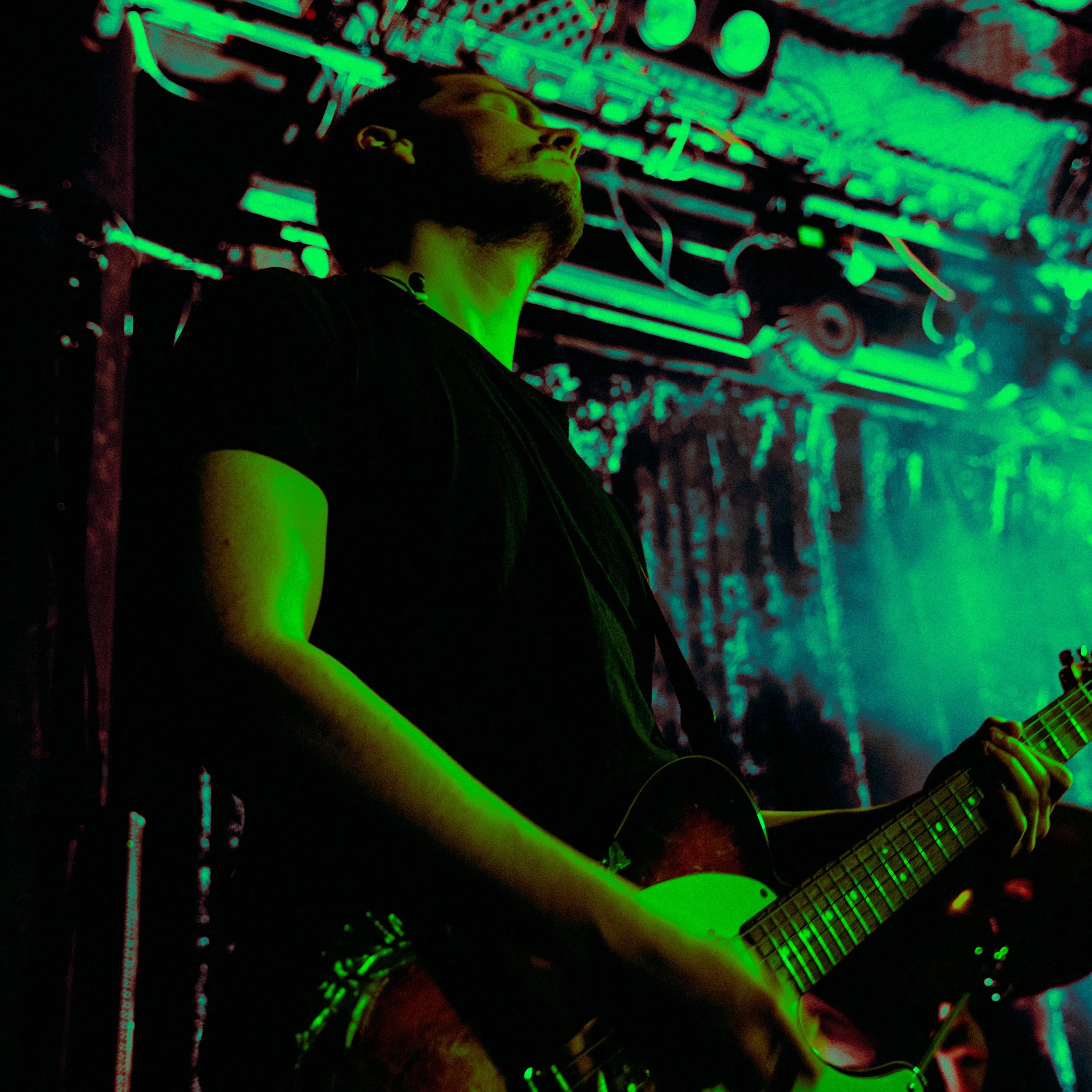 A man playing an electric guitar on stage with neon green lighting and a colorful, abstract background.
