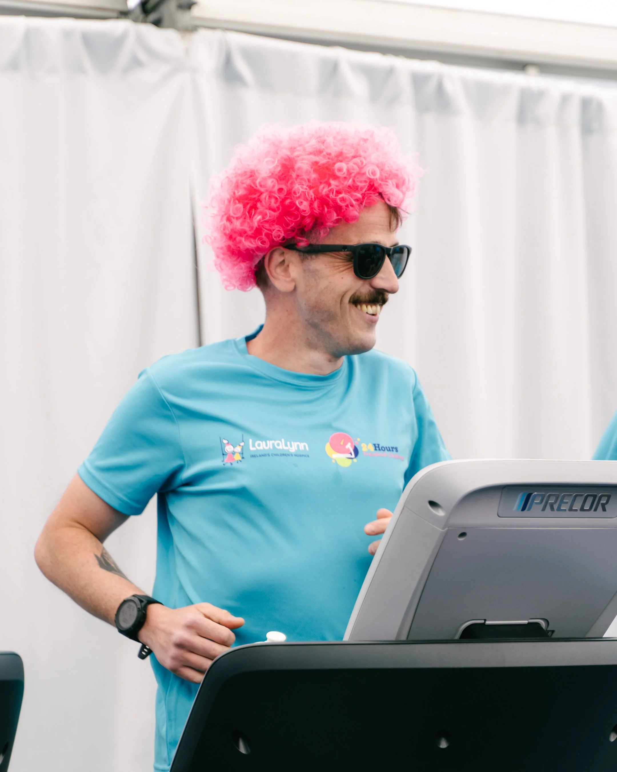 Man with a pink curly wig, sunglasses, blue t-shirt, and a wristwatch smiling while using a laptop at an event or race.