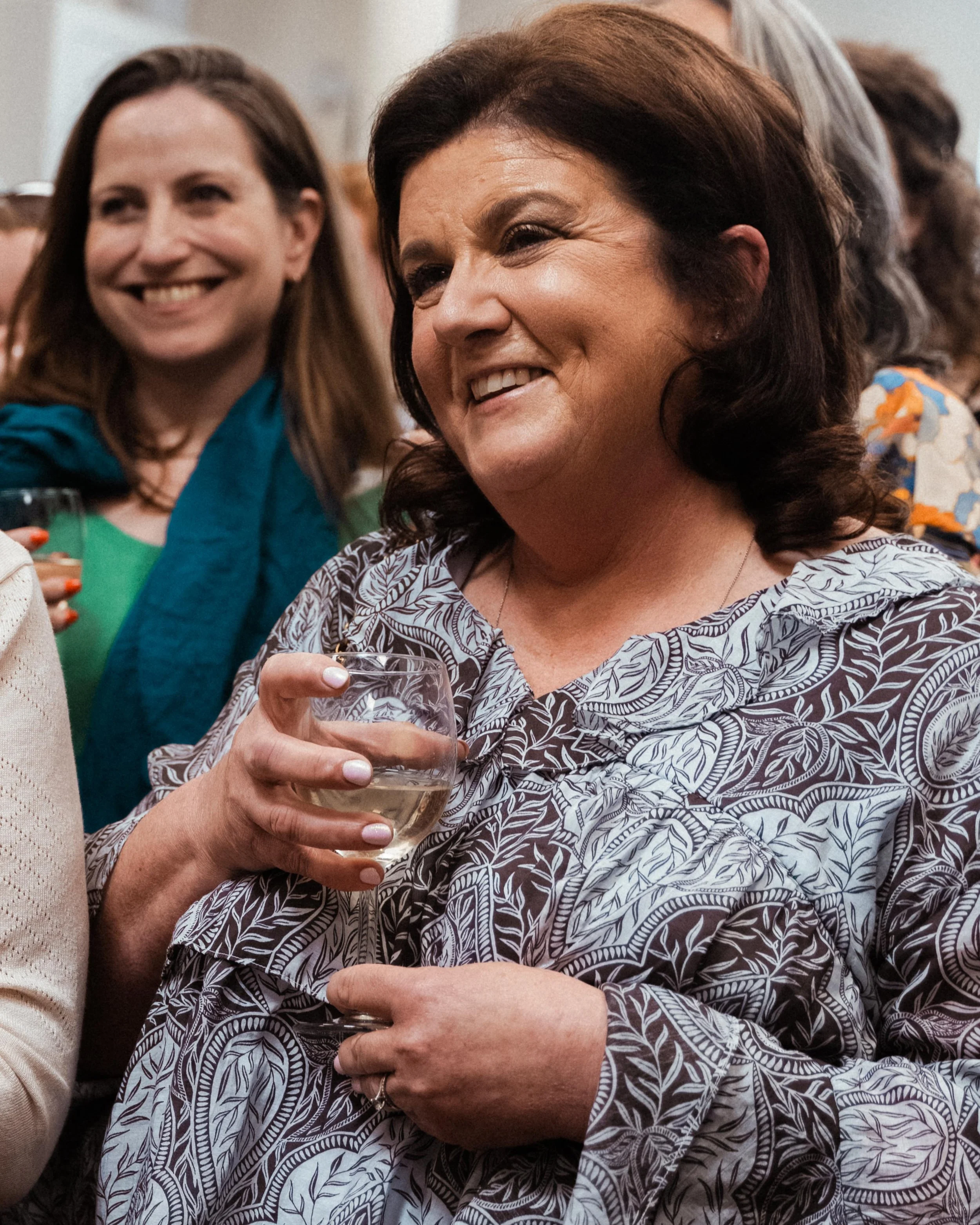 Close-up of a woman with dark hair smiling and holding a glass of white wine at a social gathering.