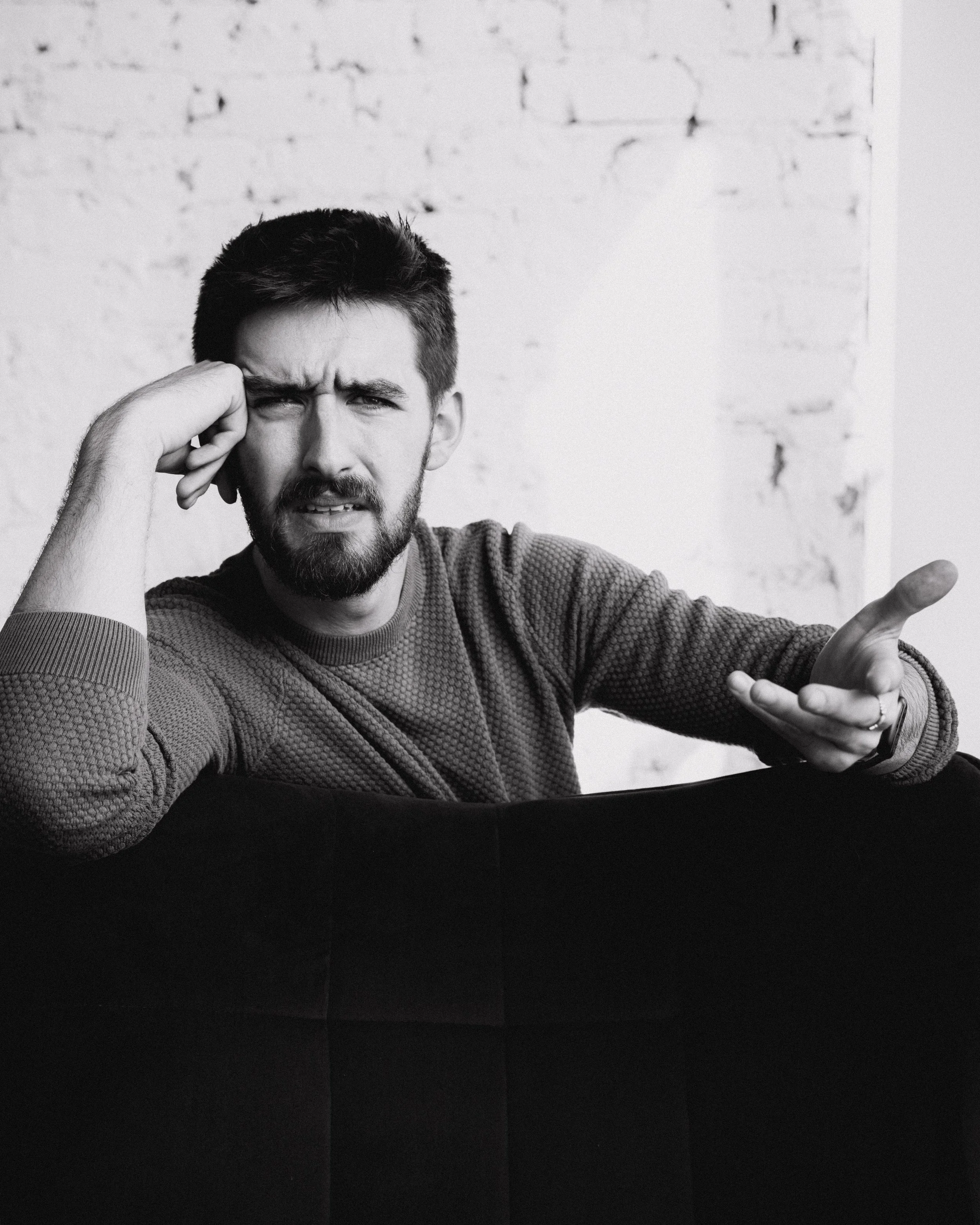 A man with a beard and short dark hair makes a confused or frustrated facial expression. He is wearing a textured, long-sleeved sweater and is gesturing with his right hand while holding his head with his left hand against a plain, light-colored bric