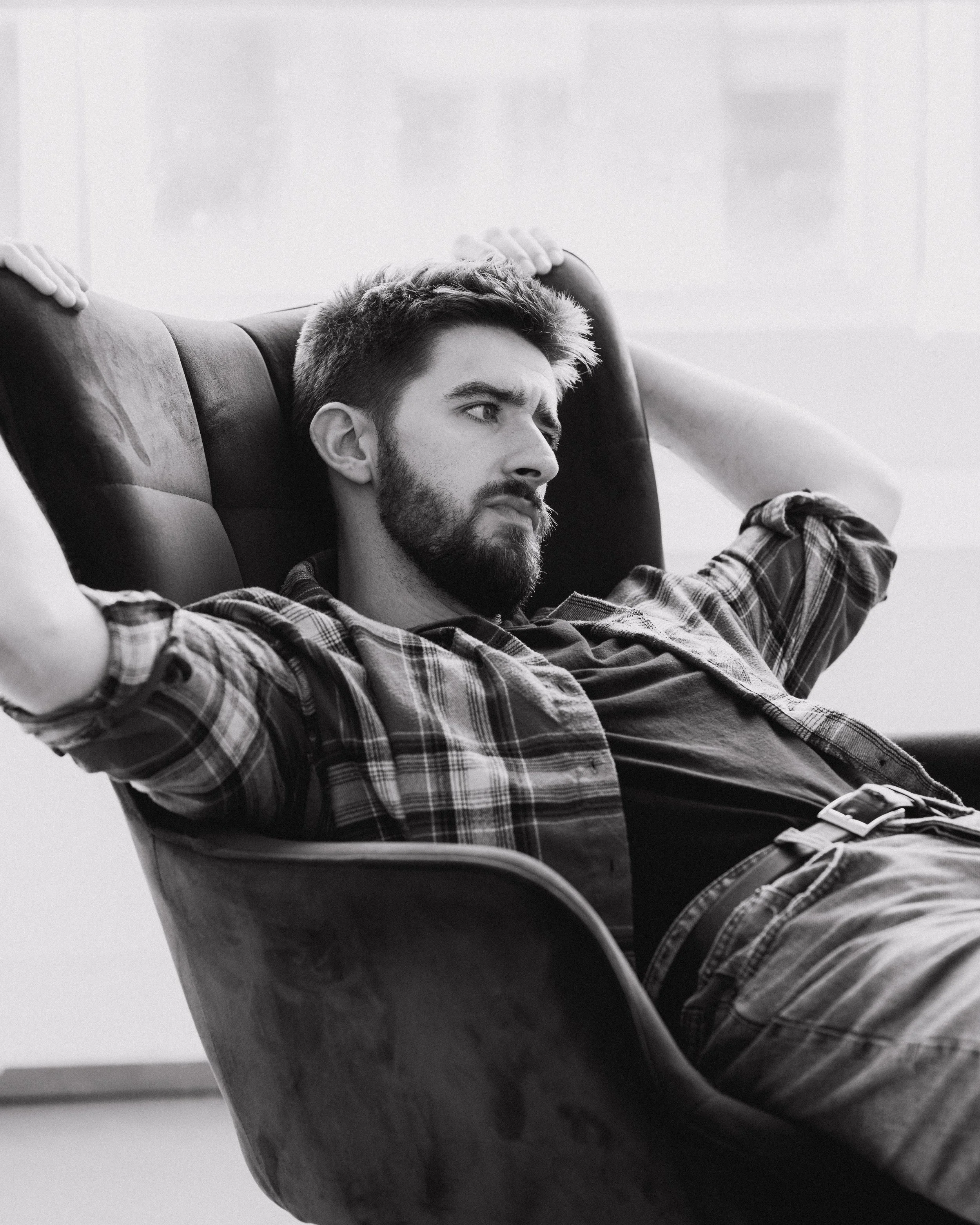 A young man with a beard and short hair, wearing a checkered shirt, is sitting in a chair with a padded backrest. He has a serious or contemplative expression, with one hand resting on his head and the other on the armrest, looking to the side. The p