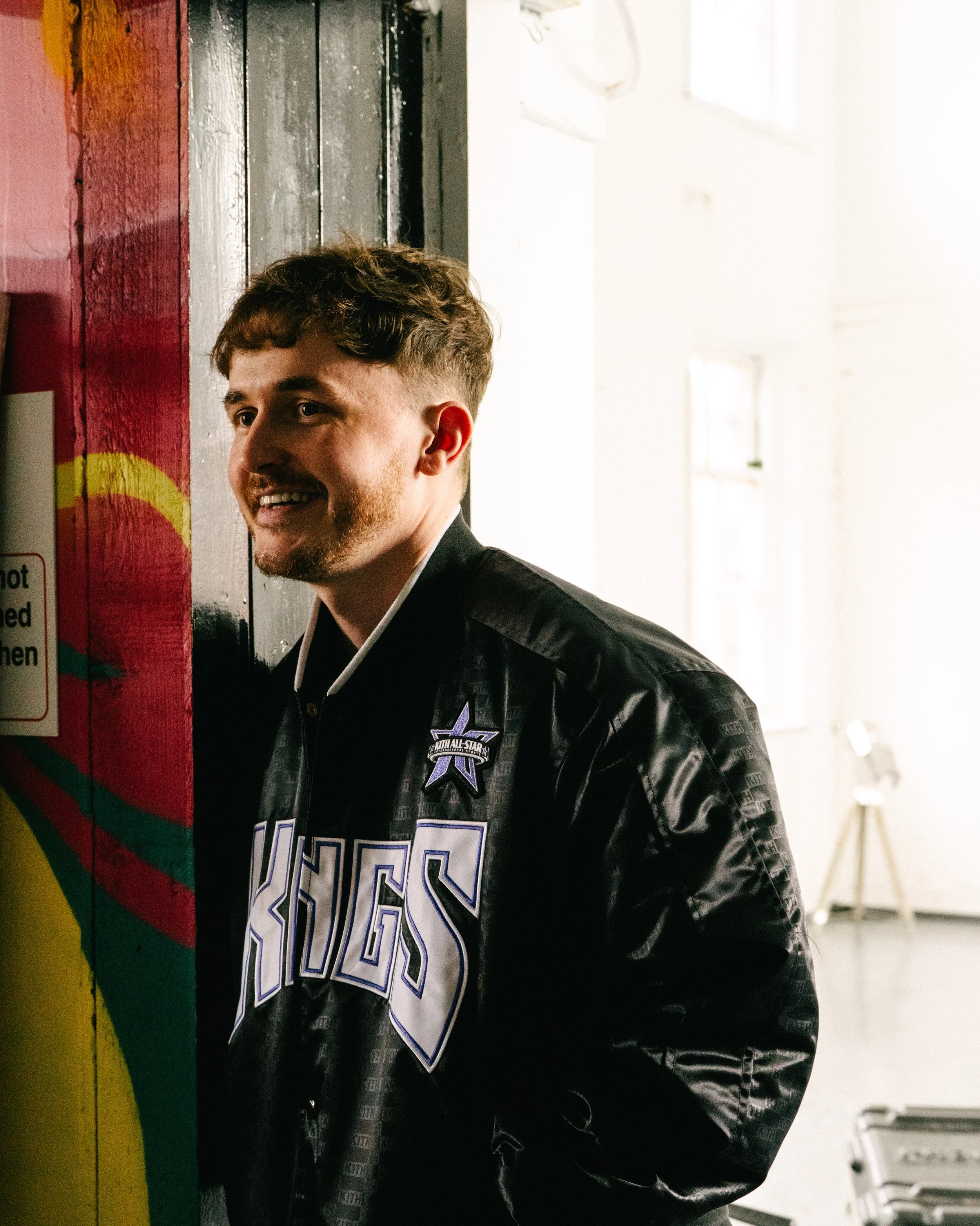A young man with short brown hair and a beard, wearing a black jacket with the Dallas Stars logo, standing near a black and red wall in a brightly lit room.