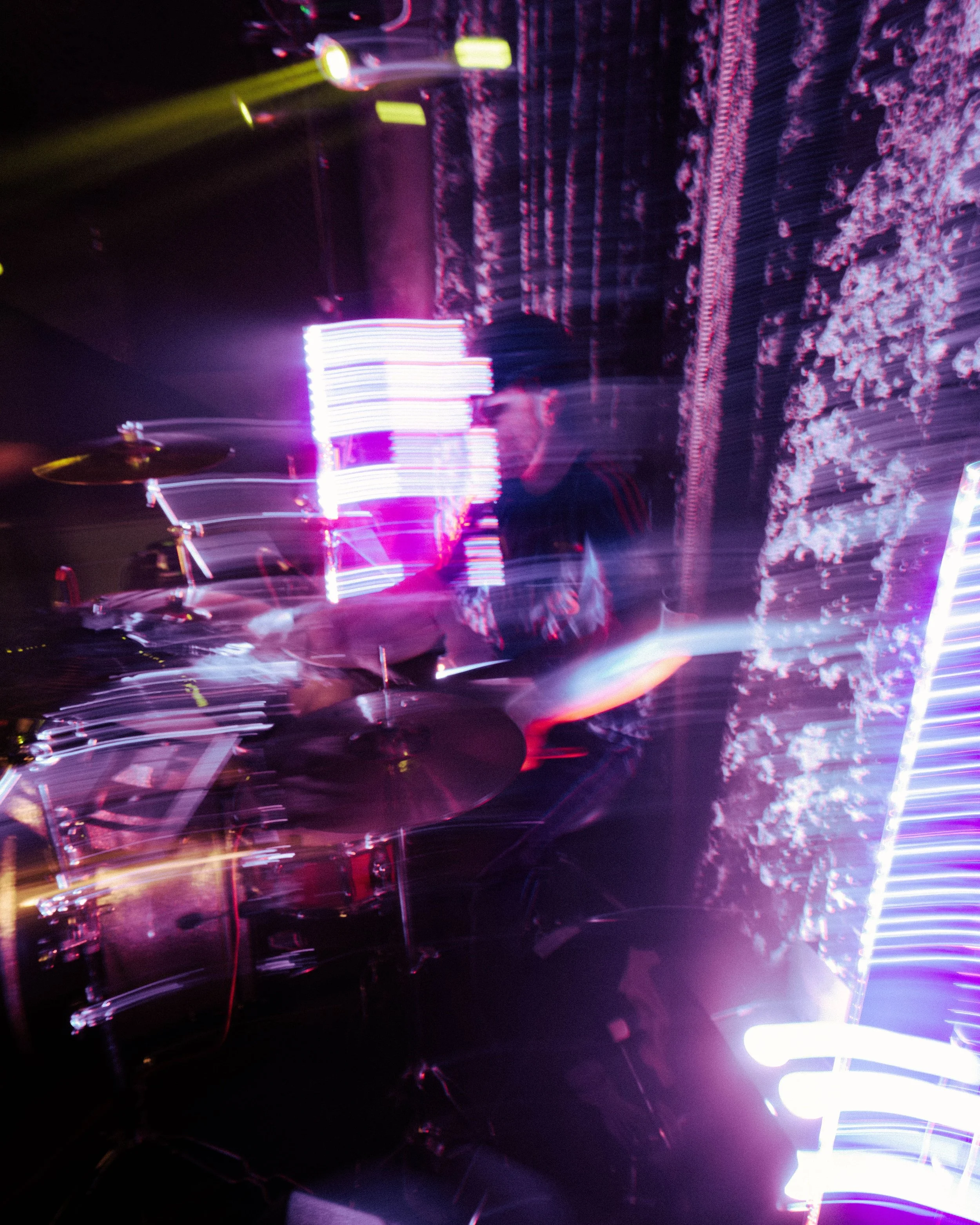 A musician playing drums in a vibrant, colorful nightclub with dynamic, blurry light effects and LED displays.