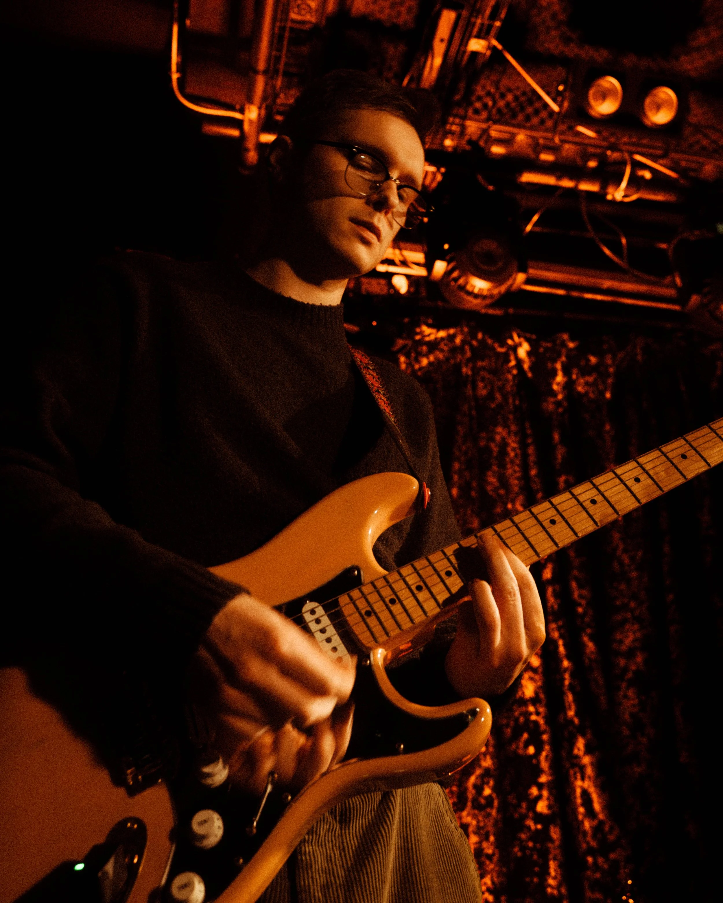 A young man with glasses playing an electric guitar on stage with red lighting and a patterned curtain in the background.