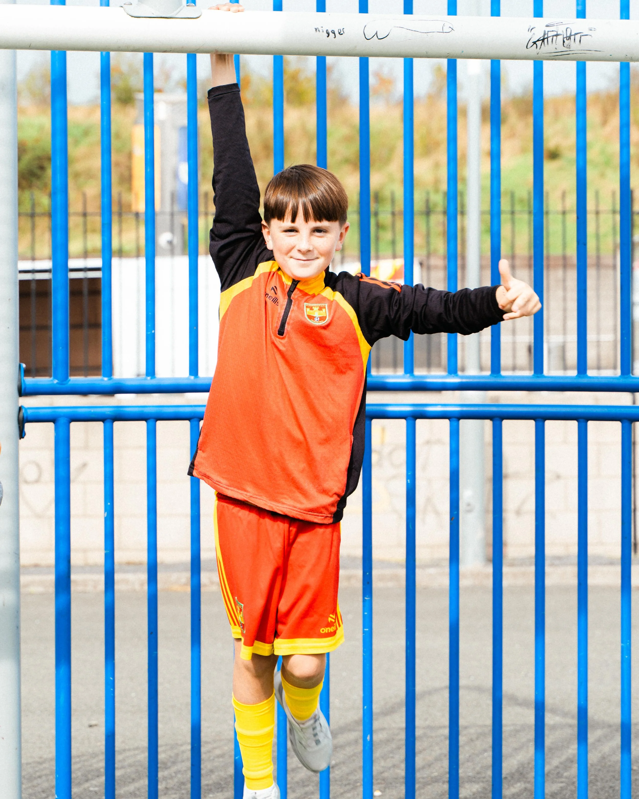 A young boy in a soccer uniform standing behind a blue metal fence, holding onto the top horizontal bar with one hand, giving a thumbs-up with his other hand, and wearing a red and yellow soccer jersey with matching shorts and knee-high socks.