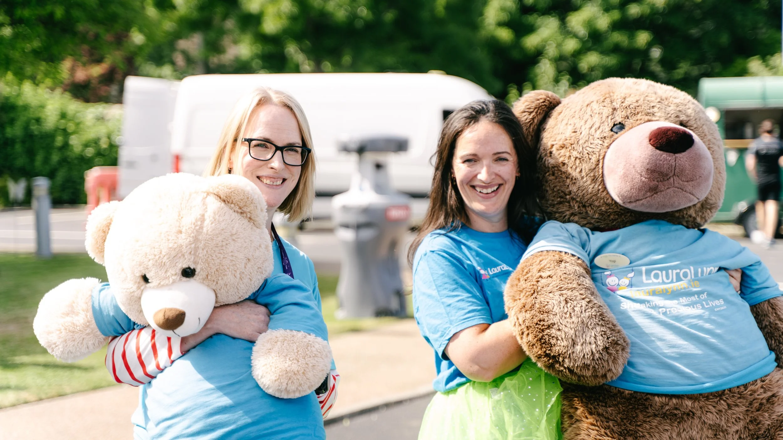 Two women smiling at the camera, each holding a large teddy bear, outdoors in a park or event setting. Both are wearing light blue T-shirts, and one woman wears glasses. There are trees and a parking lot with vehicles in the background.