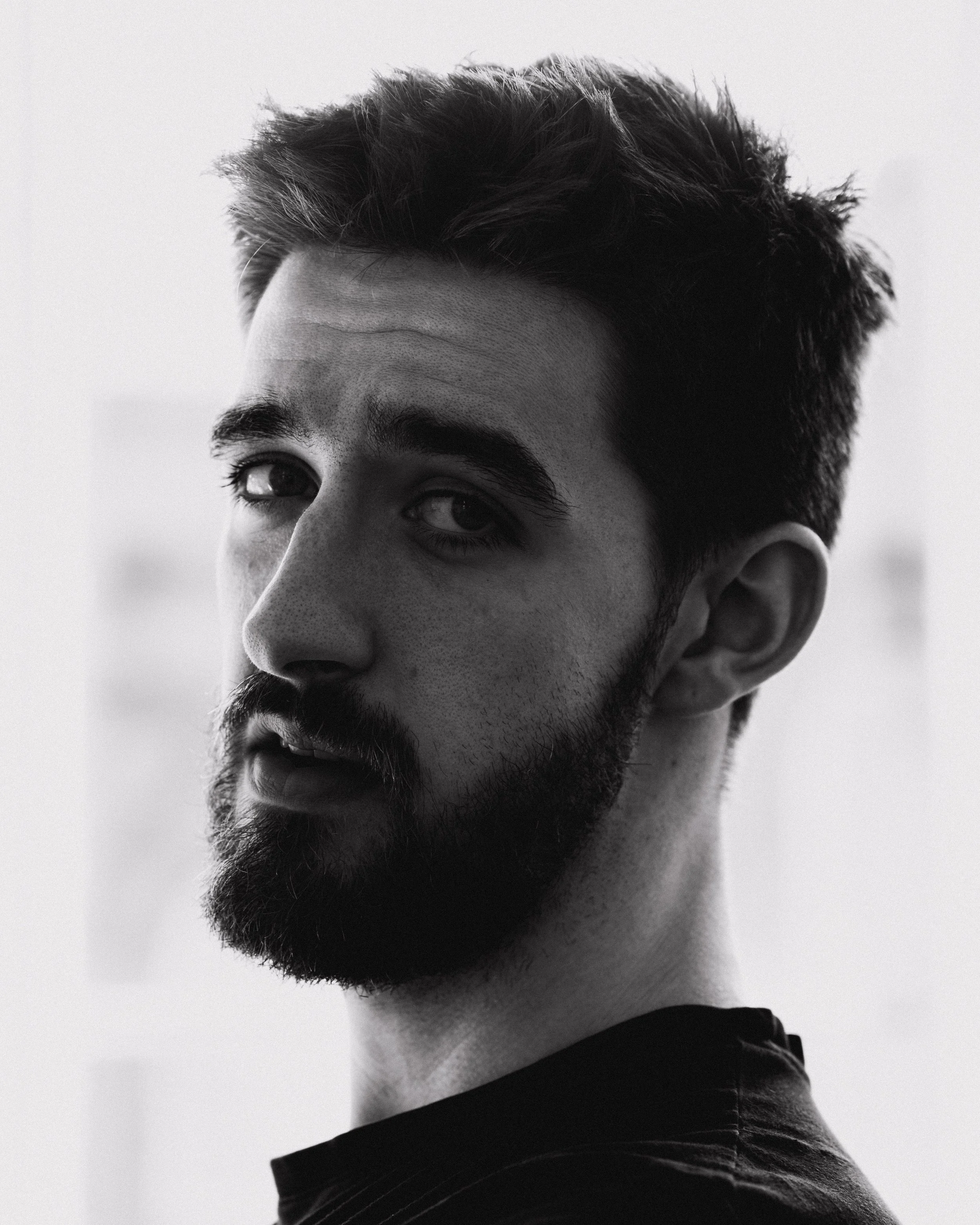 Black and white close-up photo of a young man with short, dark hair and a beard, looking slightly to the side against a plain light background.