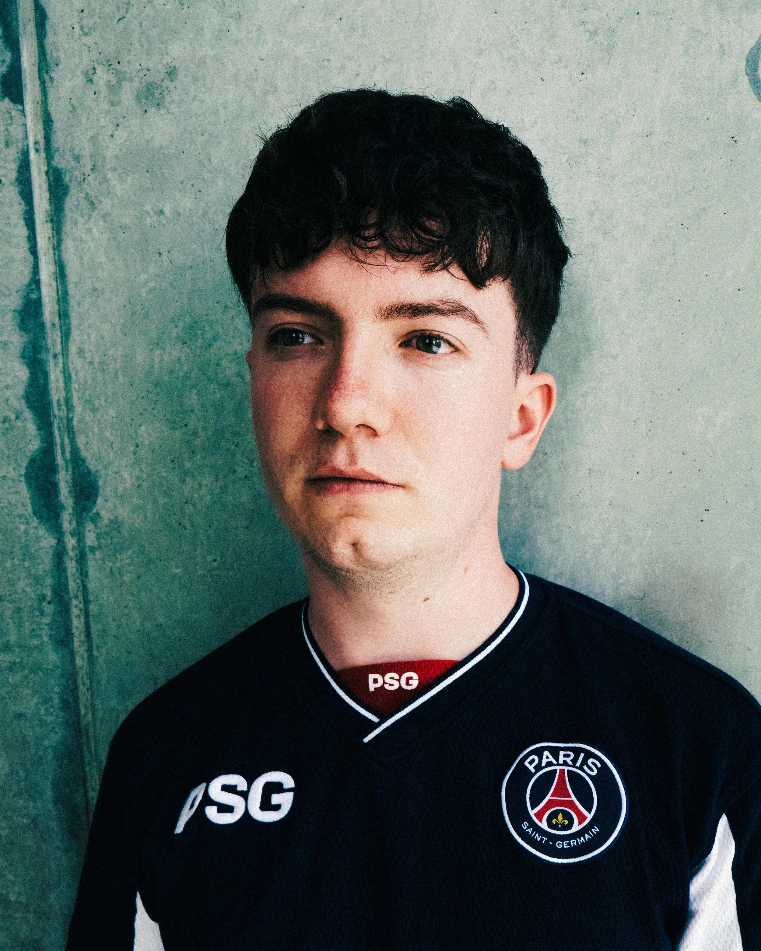 Close-up of a young man with dark curly hair, wearing a black Paris Saint-Germain football club jersey with the PSG logo on the chest, standing against a concrete wall.