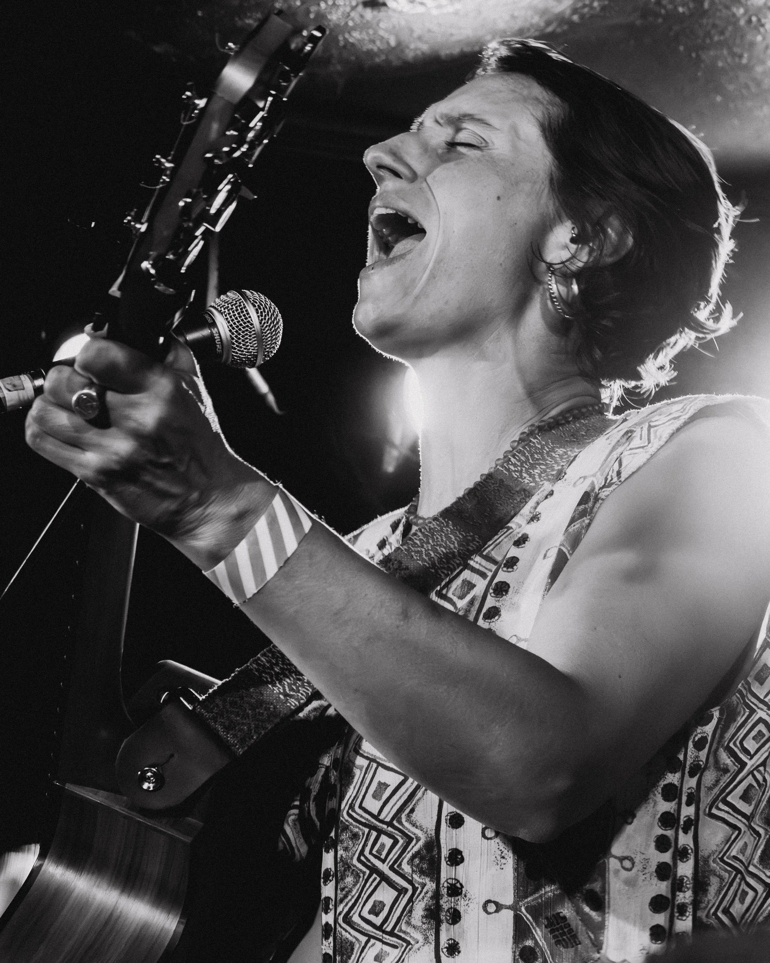 A woman with short, dark hair singing passionately into a microphone on stage. She is dressed in traditional clothing with decorative patterns and jewelry. The photo is in black and white and shows a cultural performance setting.