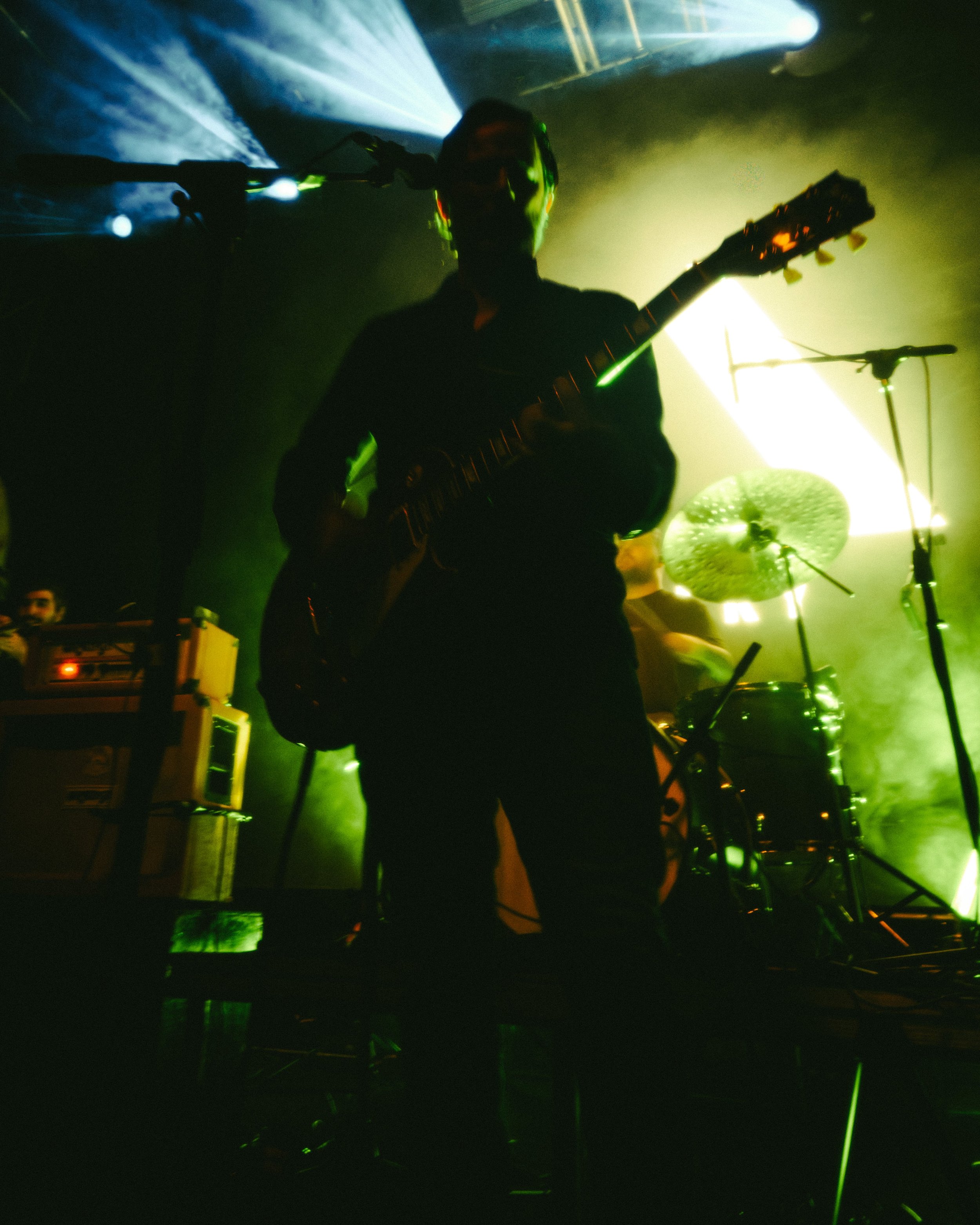 A musician playing an electric guitar on stage with vibrant green and yellow stage lighting and a drum set in the background.