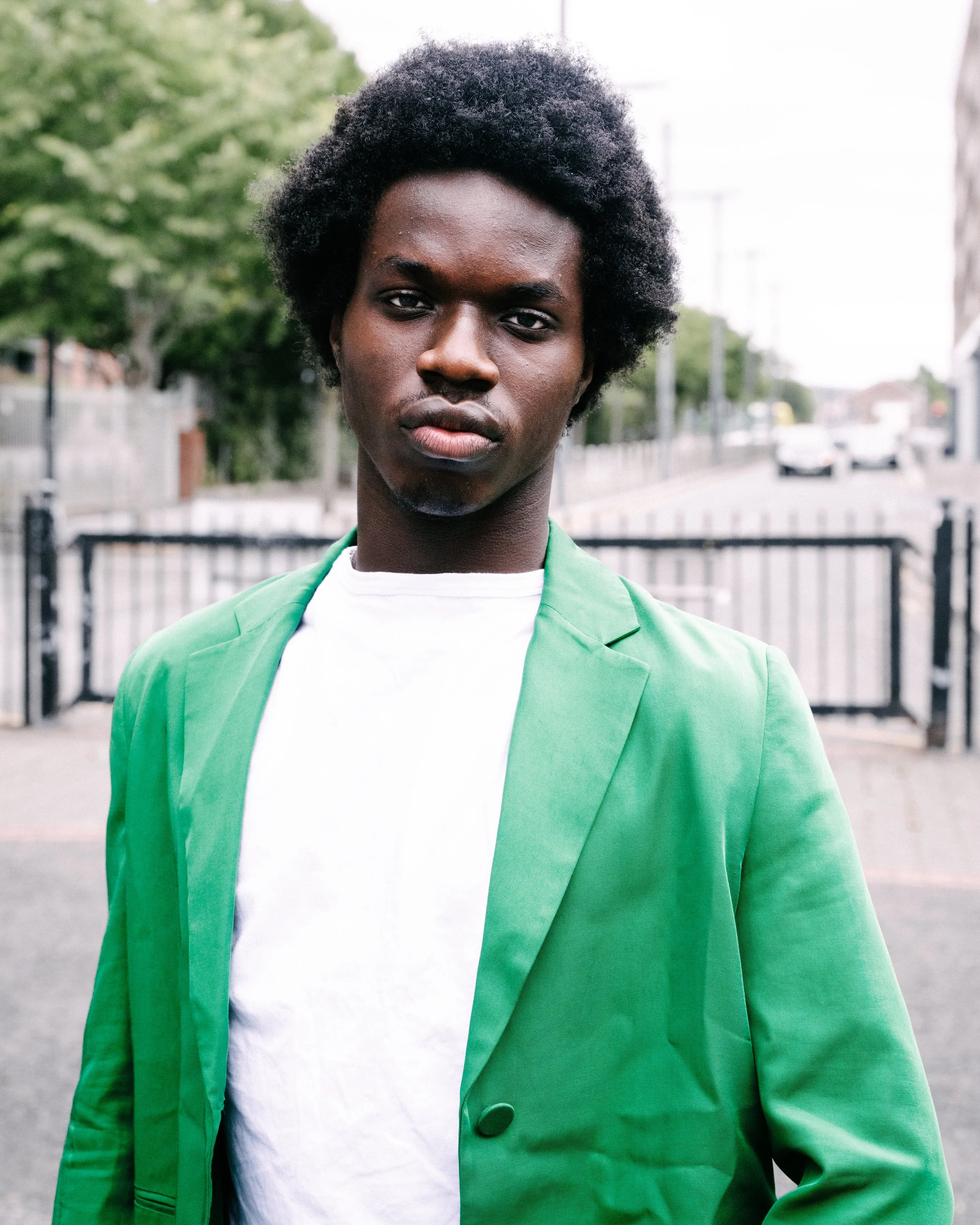 A young person with short curly hair wearing a green jacket and white shirt, standing outdoors with trees, a fence, and a street with cars in the background.