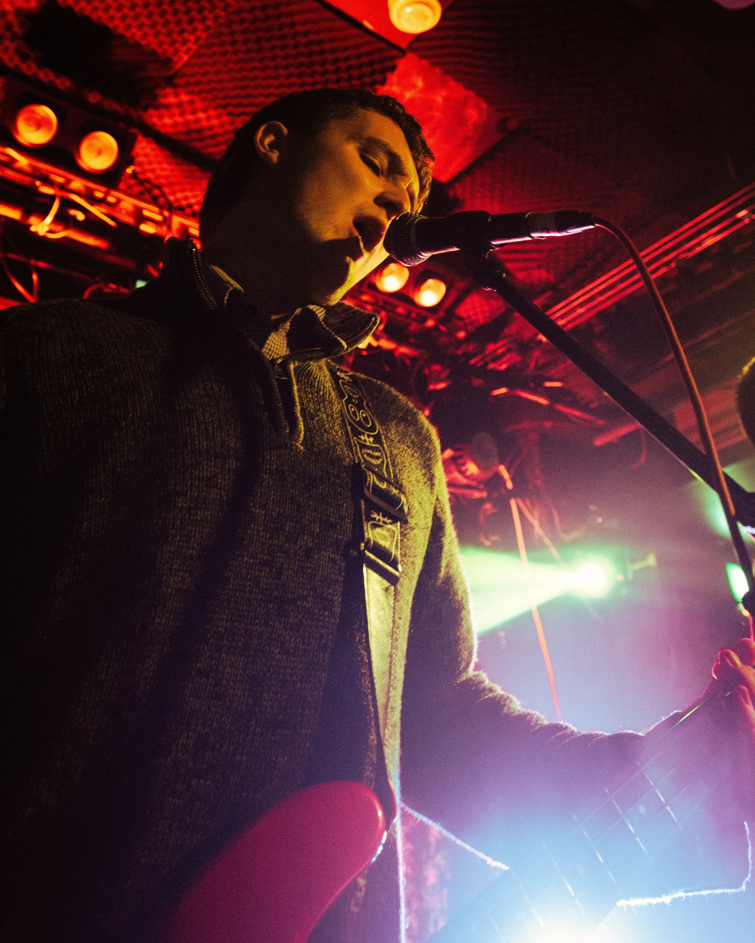 A person singing into a microphone with a guitar, illuminated by colorful stage lights, in a dimly lit club or concert venue.