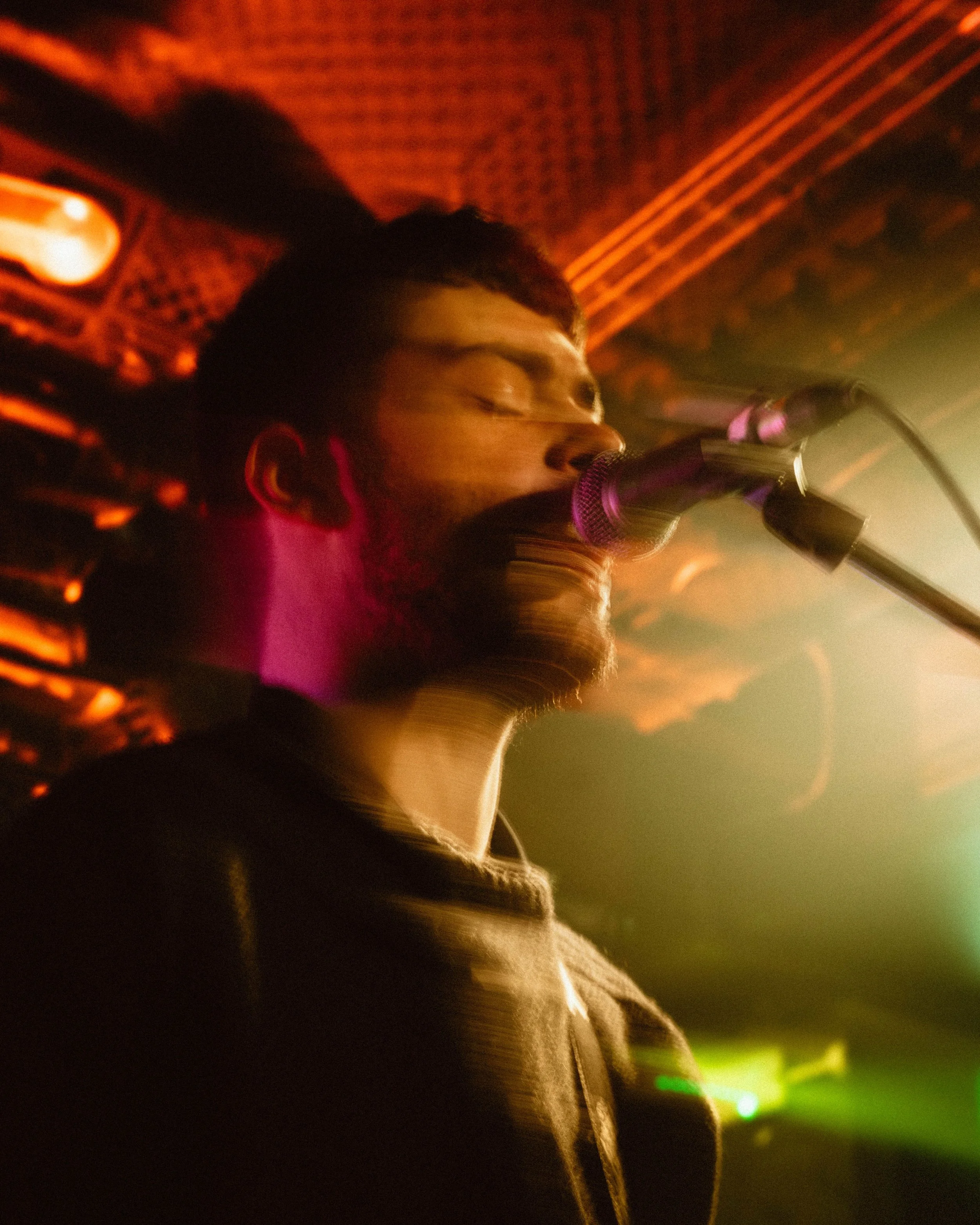 A male singer closes his eyes while singing into a microphone, illuminated by colorful stage lights in a dim, atmospheric setting.