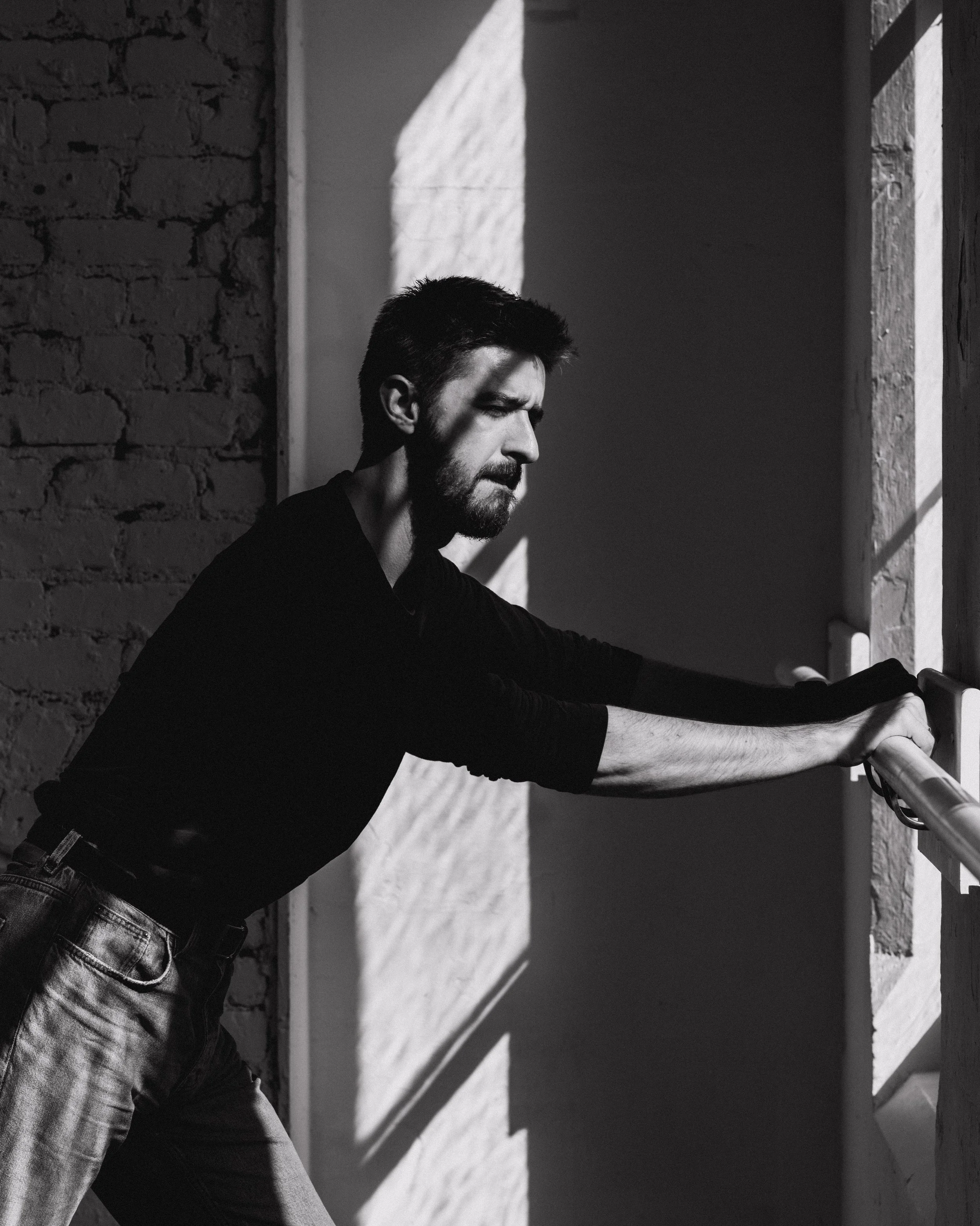 A black-and-white photo of a man with short dark hair and a beard, wearing a dark long-sleeve shirt and jeans, sitting and stretching his arms forward in front of a textured brick wall with sunlight casting shadows.