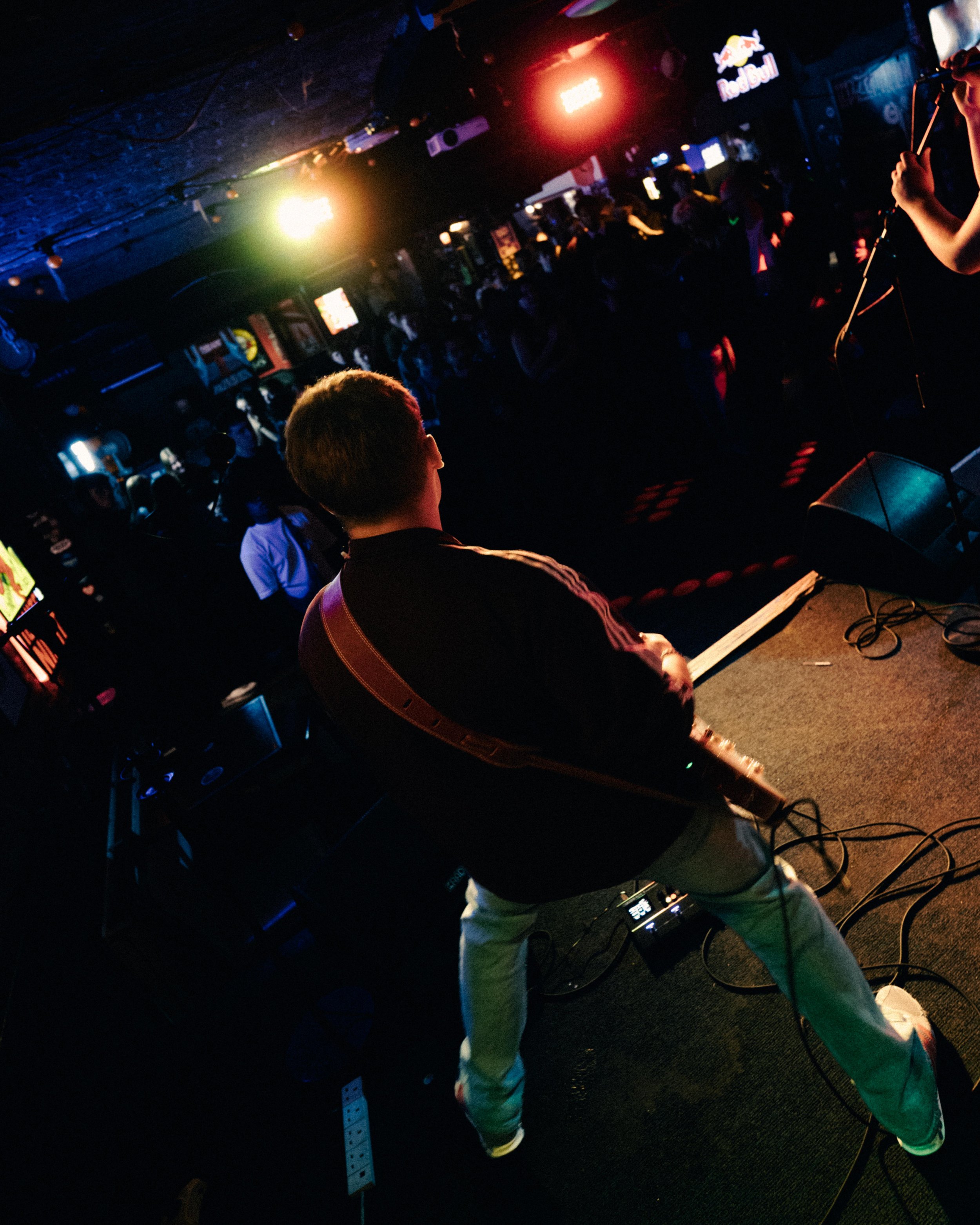 A person playing an electronic drum pad in a dark nightclub or bar with colorful lights and a crowd of people in the background.