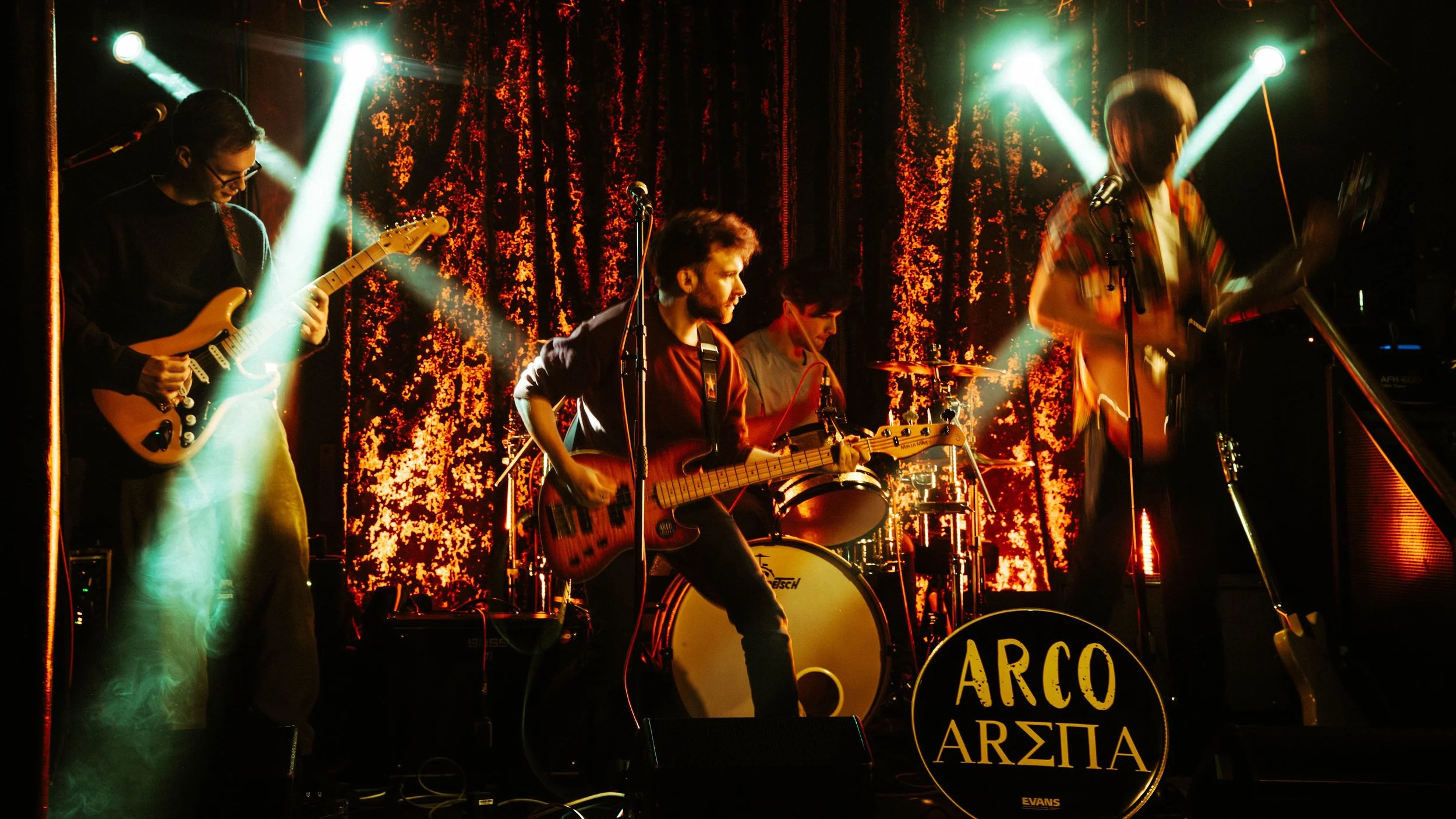 A band performing live on stage with four musicians, colorful lighting, and a textured red and orange backdrop.
