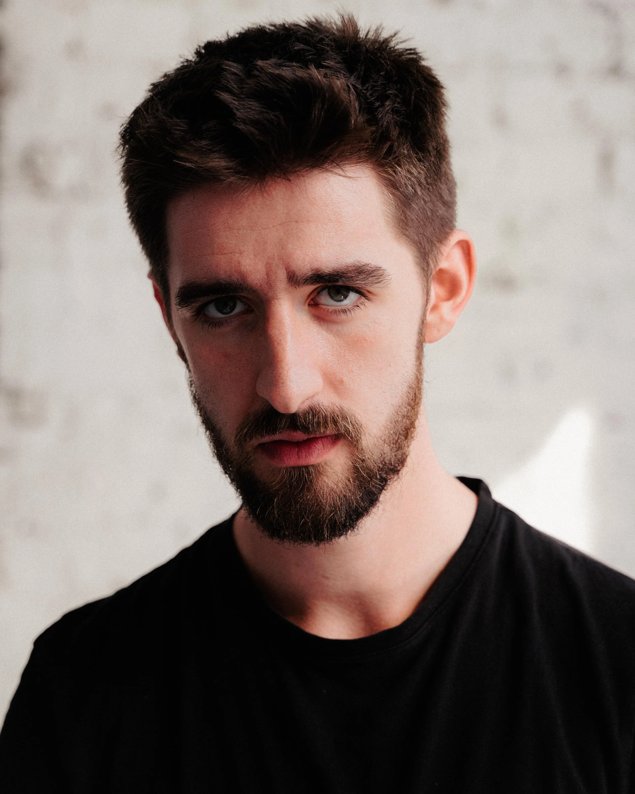 Close-up of a man with dark hair and a beard wearing a black shirt, looking directly at the camera, against a plain light-colored background.